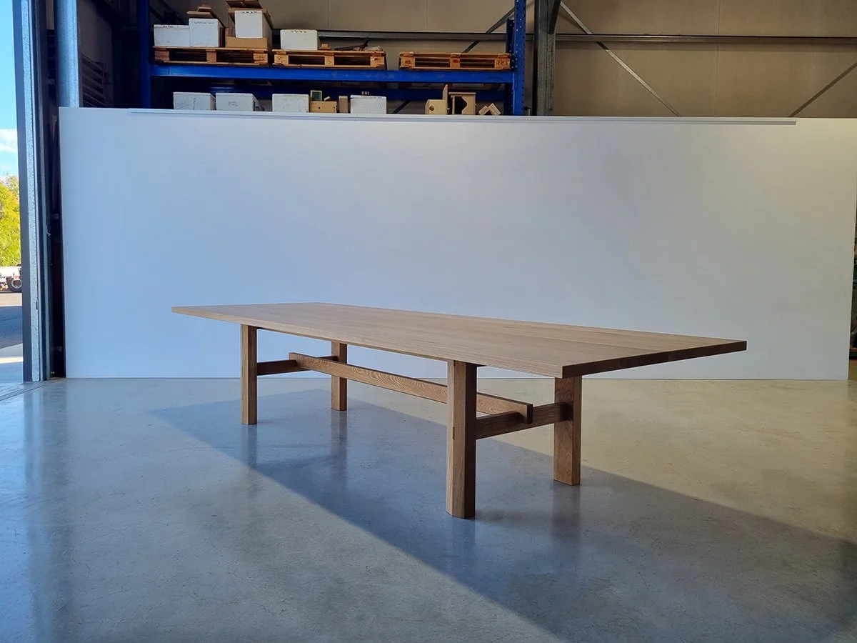 Laura's Table, a 12-seat farmhouse-style dining table in French Oak with exposed joinery, front-on view in workshop setting - bespoke furniture by Nathan Day Design