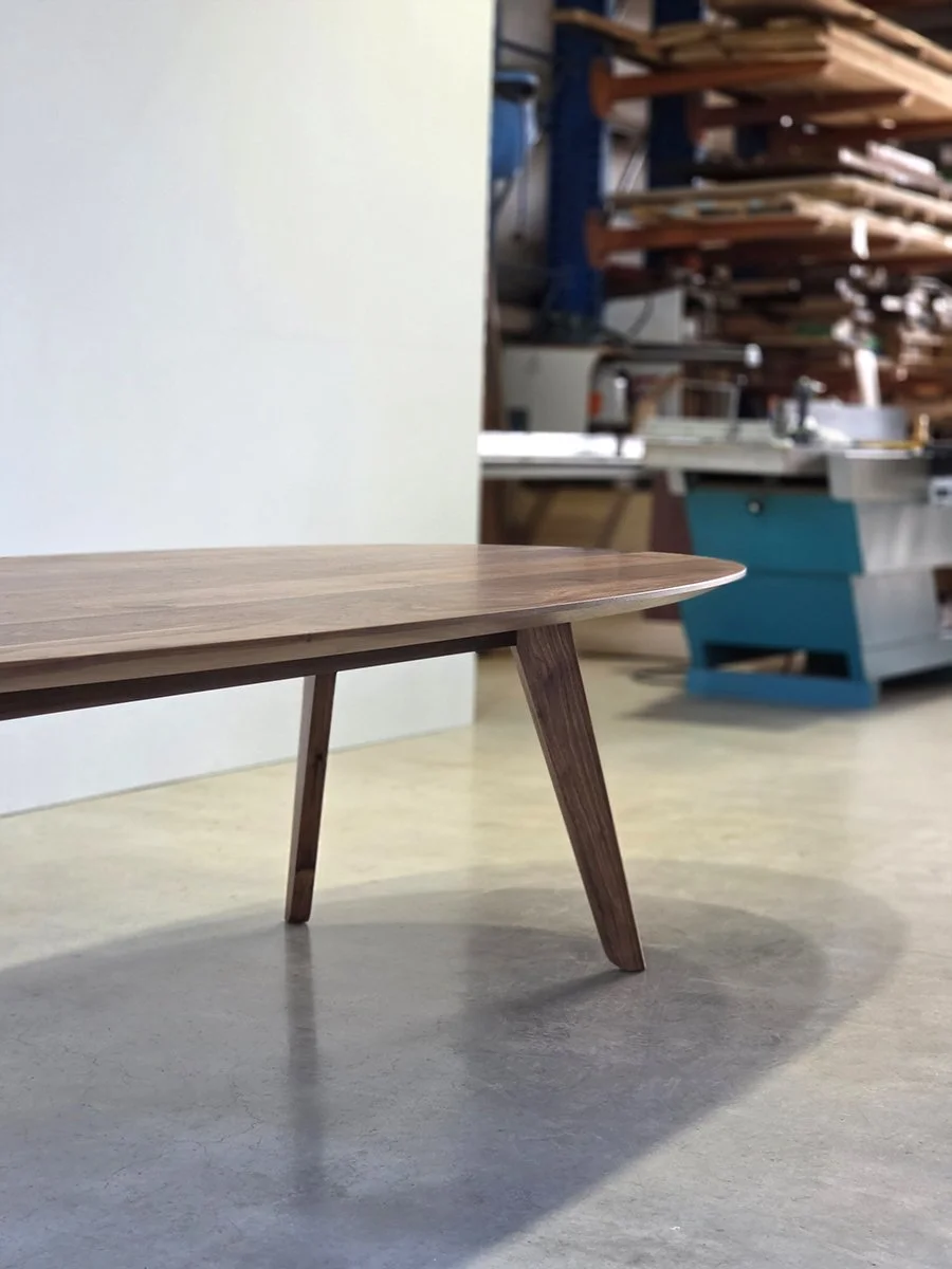 Angled view of Peppy Grove Table in Walnut showing tapered leg detail, workshop setting with machinery in background - bespoke furniture by Nathan Day Design