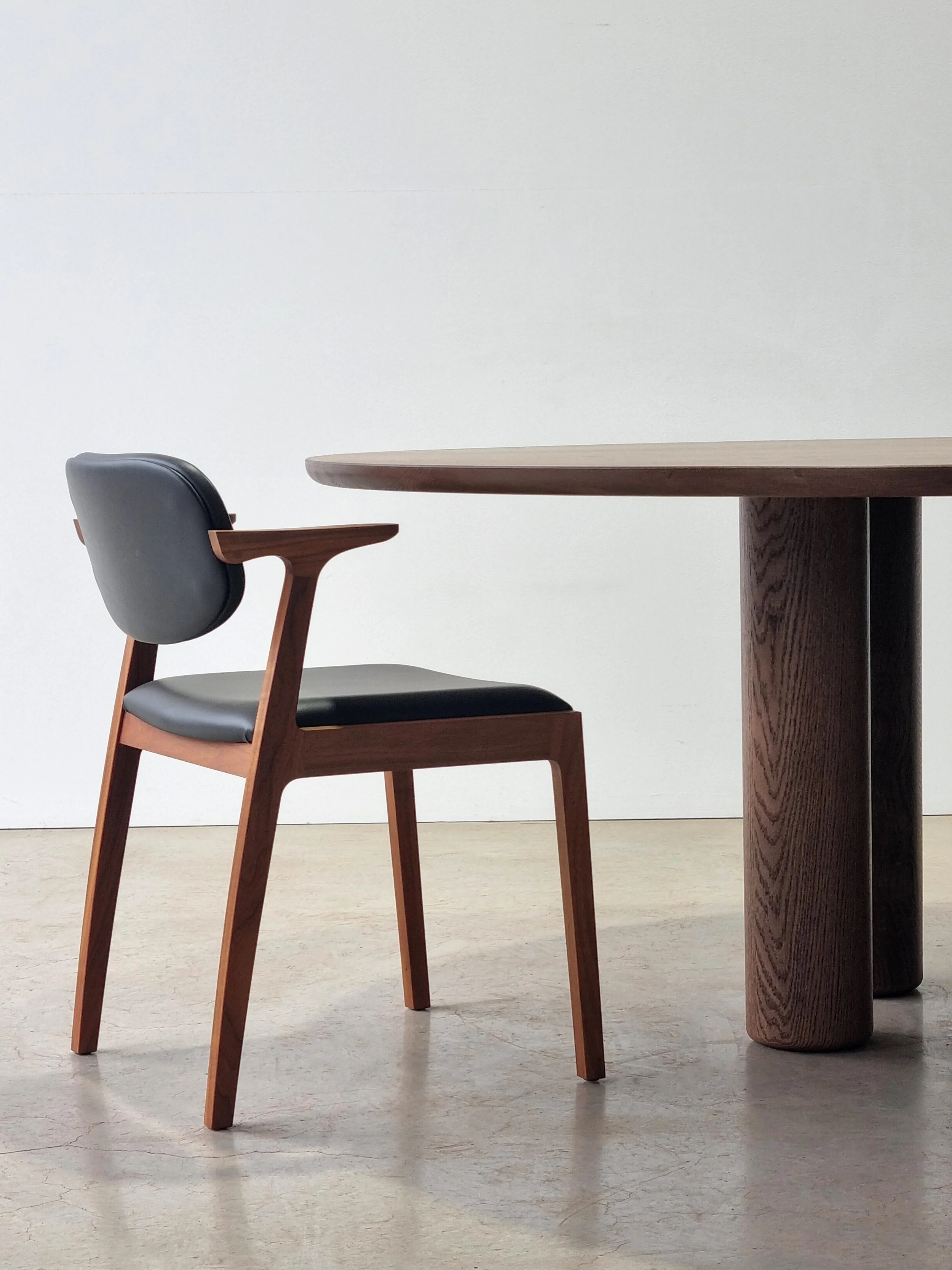 Dark timber-armed dining chair with black leather upholstery beside the Anemone Table's tapered column leg - Nathan Day Design