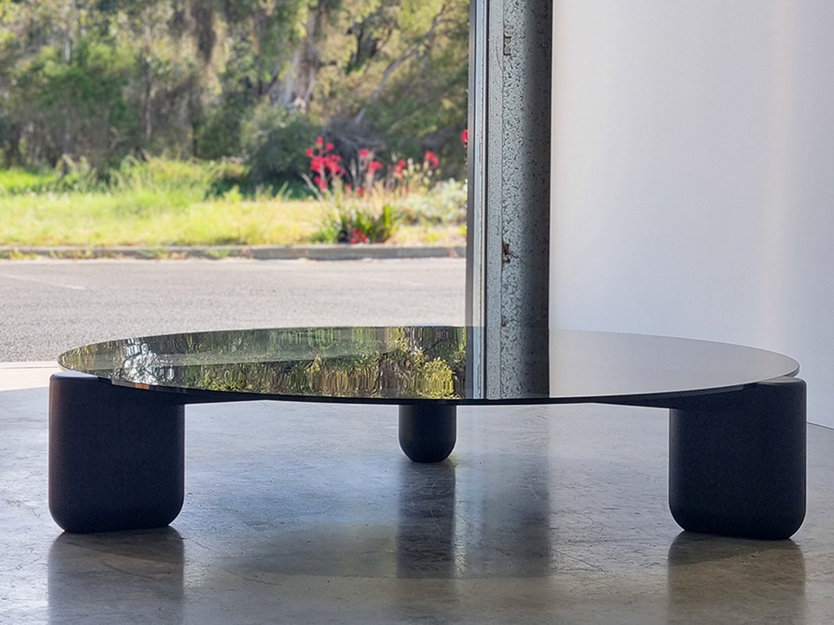 Modern round glass-top coffee table with black legs by Nathan Day Design, set beside a window overlooking greenery.