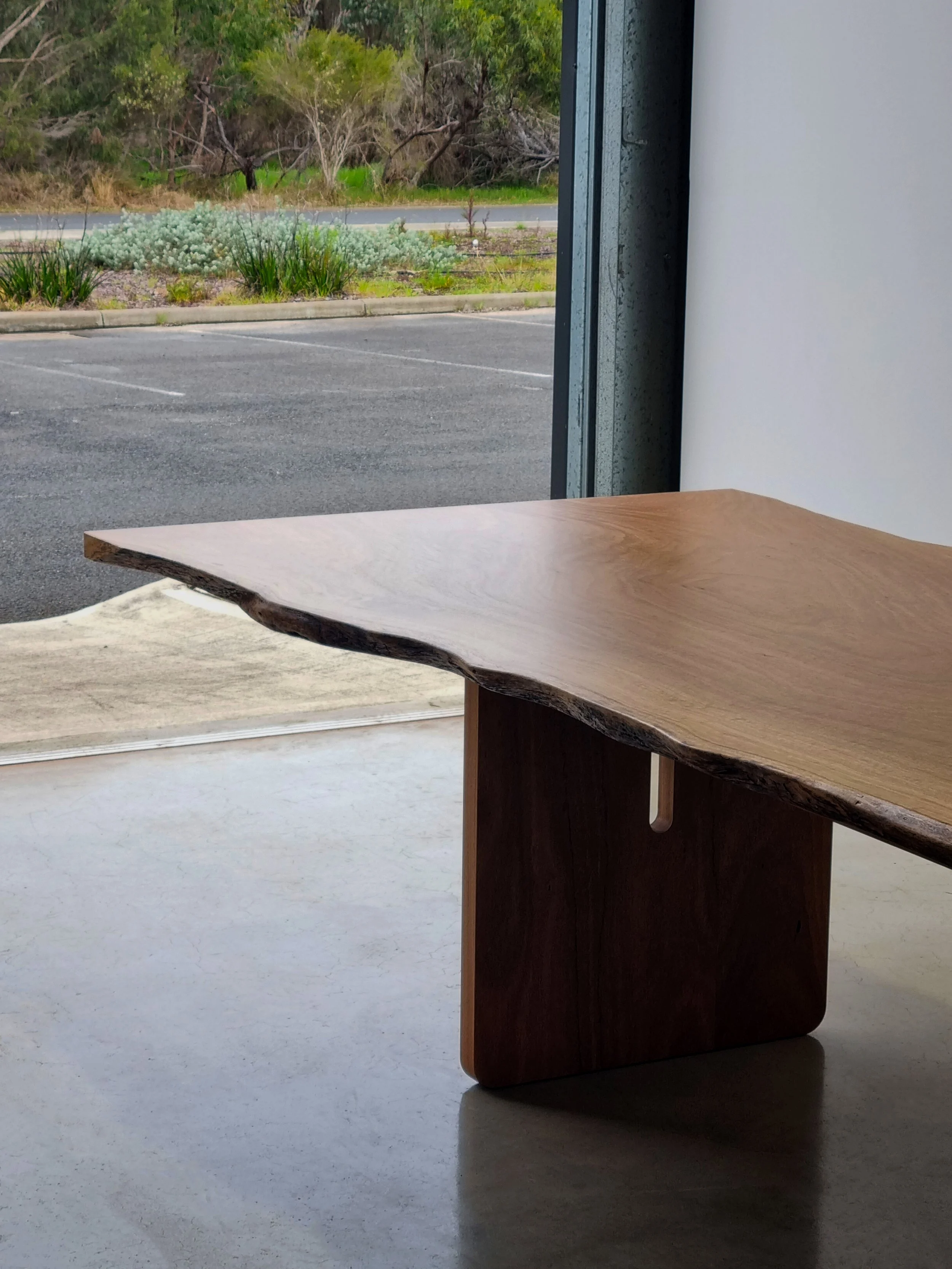 Live edge dining table with slotted timber leg detail near open workshop roller door, showing the natural undulating edge - bespoke furniture by Nathan Day Design
