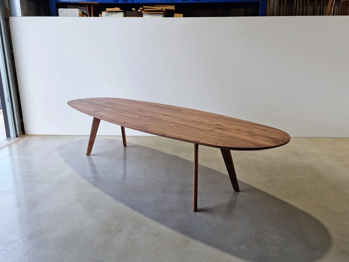 Full angled view of Peppy Grove Table in Walnut showing its surfboard-like oval form and tapered legs, near workshop doorway - bespoke furniture by Nathan Day Design