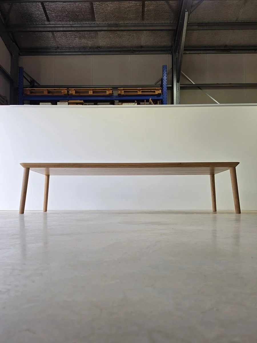 Low-angle view of Mercator Way Table in American Oak showing tapered legs and slim profile, workshop setting with industrial roof beams - bespoke furniture by Nathan Day Design