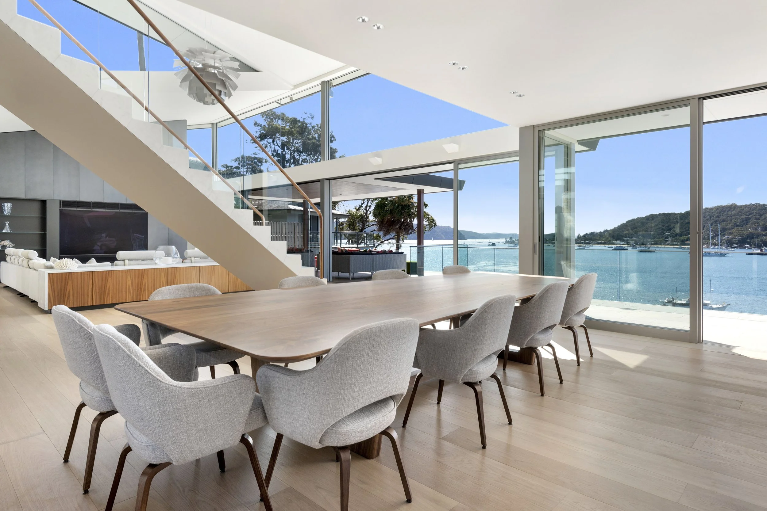 Cabarita Rd Table in Walnut set with grey upholstered chairs beside a floating staircase, expansive bay views through glass walls - bespoke furniture by Nathan Day Design