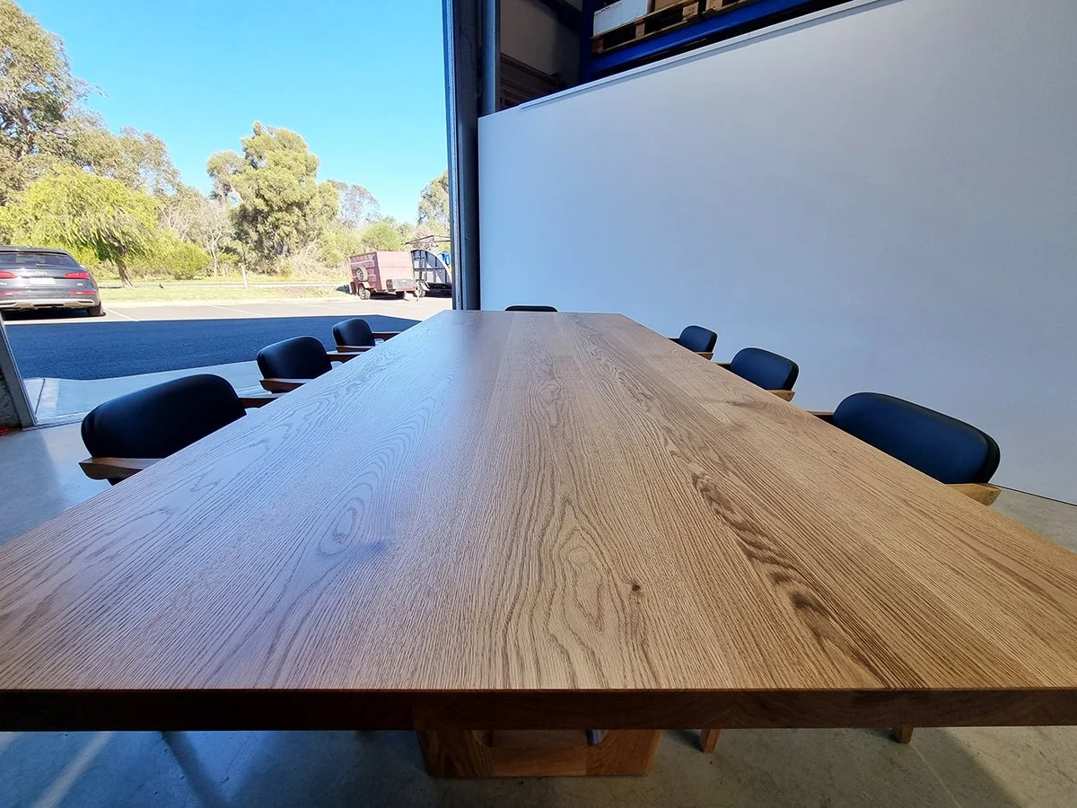 Low-angle view along Clairault Table's Oak tabletop showing rich grain, set with navy dining chairs near workshop roller door - bespoke furniture by Nathan Day Design