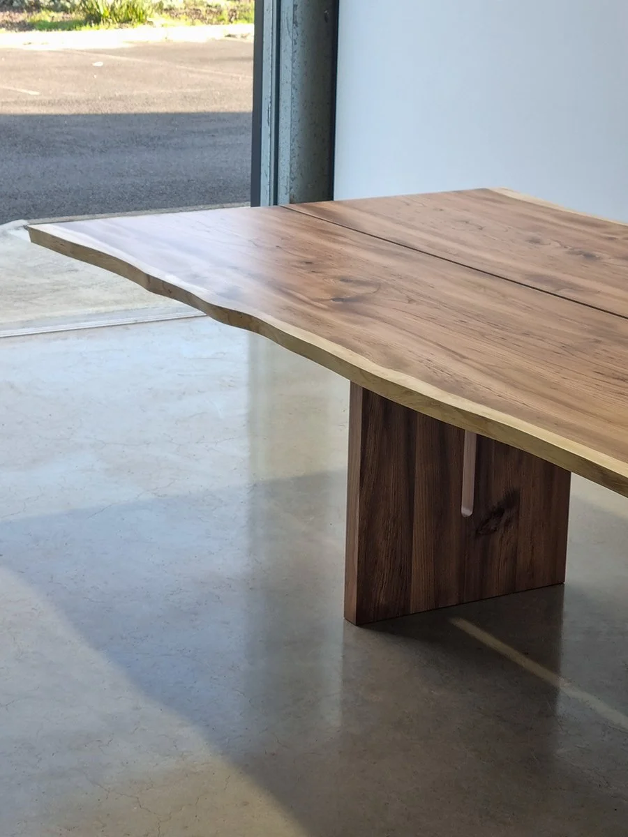Close angled view of a live edge table's joined two-slab tabletop and solid plinth leg, near workshop doorway - bespoke furniture by Nathan Day Design
