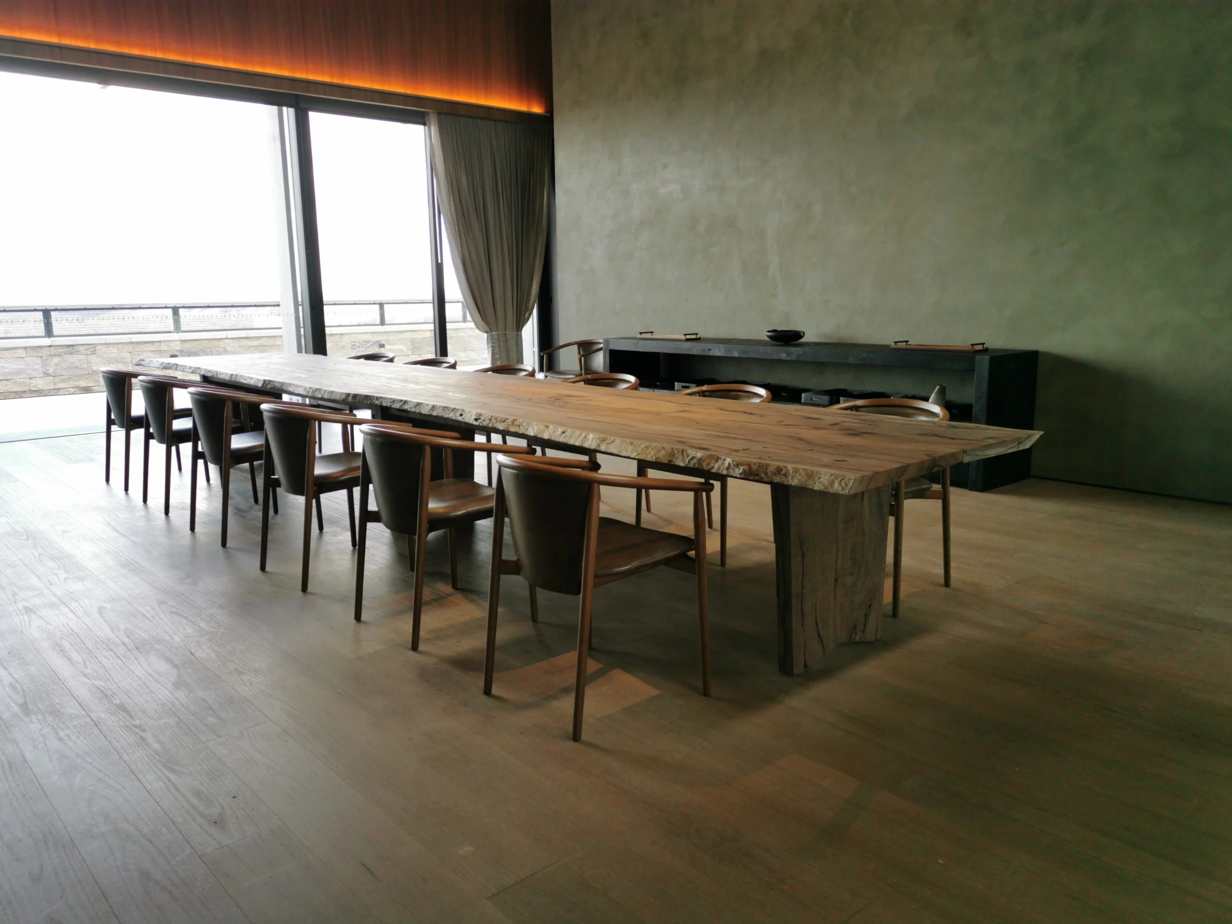Large live edge dining table set for many in an architectural dining room with floor-to-ceiling windows and moody stone walls - bespoke furniture by Nathan Day Design
