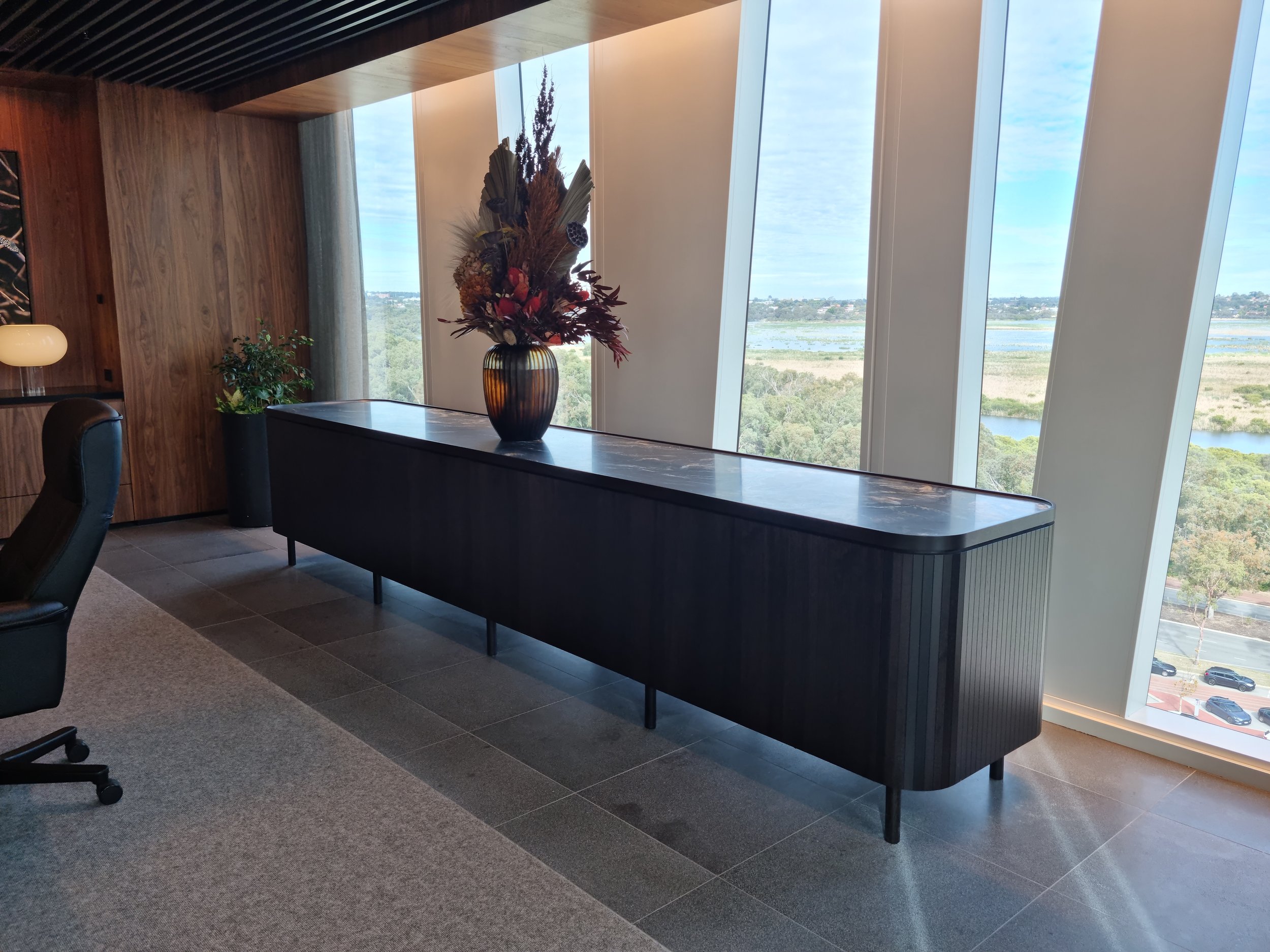 Black console table with a floral arrangement by Nathan Day Design, in a modern lobby interior with large windows overlooking greenery and water.