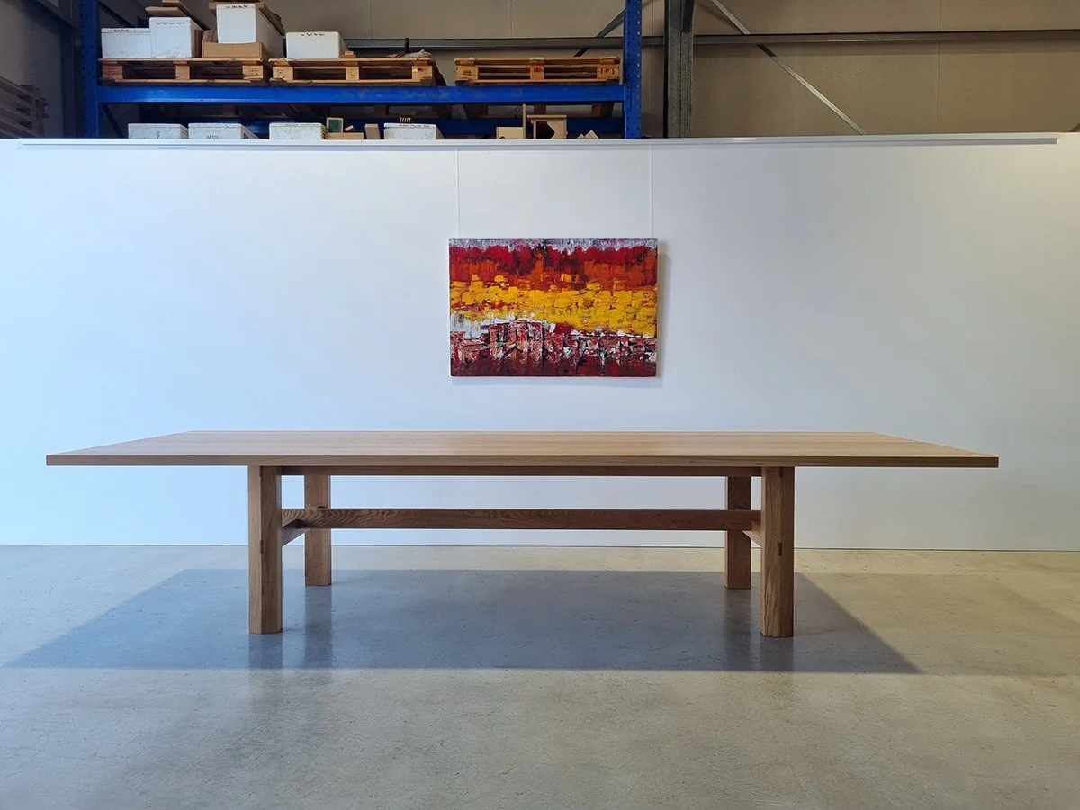 Laura's Table in French Oak with exposed stretcher joinery, workshop setting with vibrant abstract painting on the wall behind - bespoke furniture by Nathan Day Design
