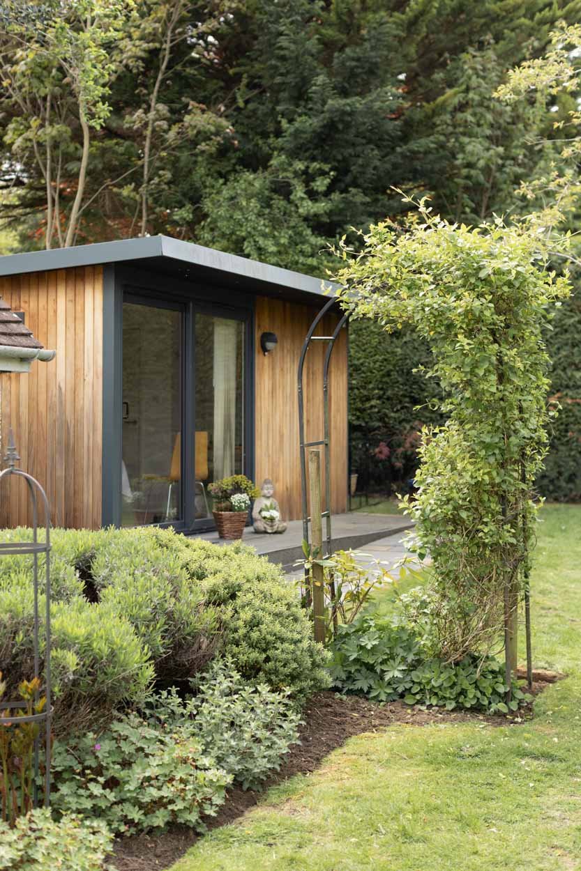 A modern wooden acupuncture treatment room with sliding glass doors, surrounded by lush greenery and a small garden with various plants and shrubs.