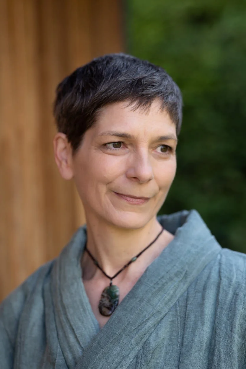 A woman with short dark hair and light skin wearing a gray top and a necklace with a large stone pendant, standing outdoors near a wooden structure with green foliage in the background.