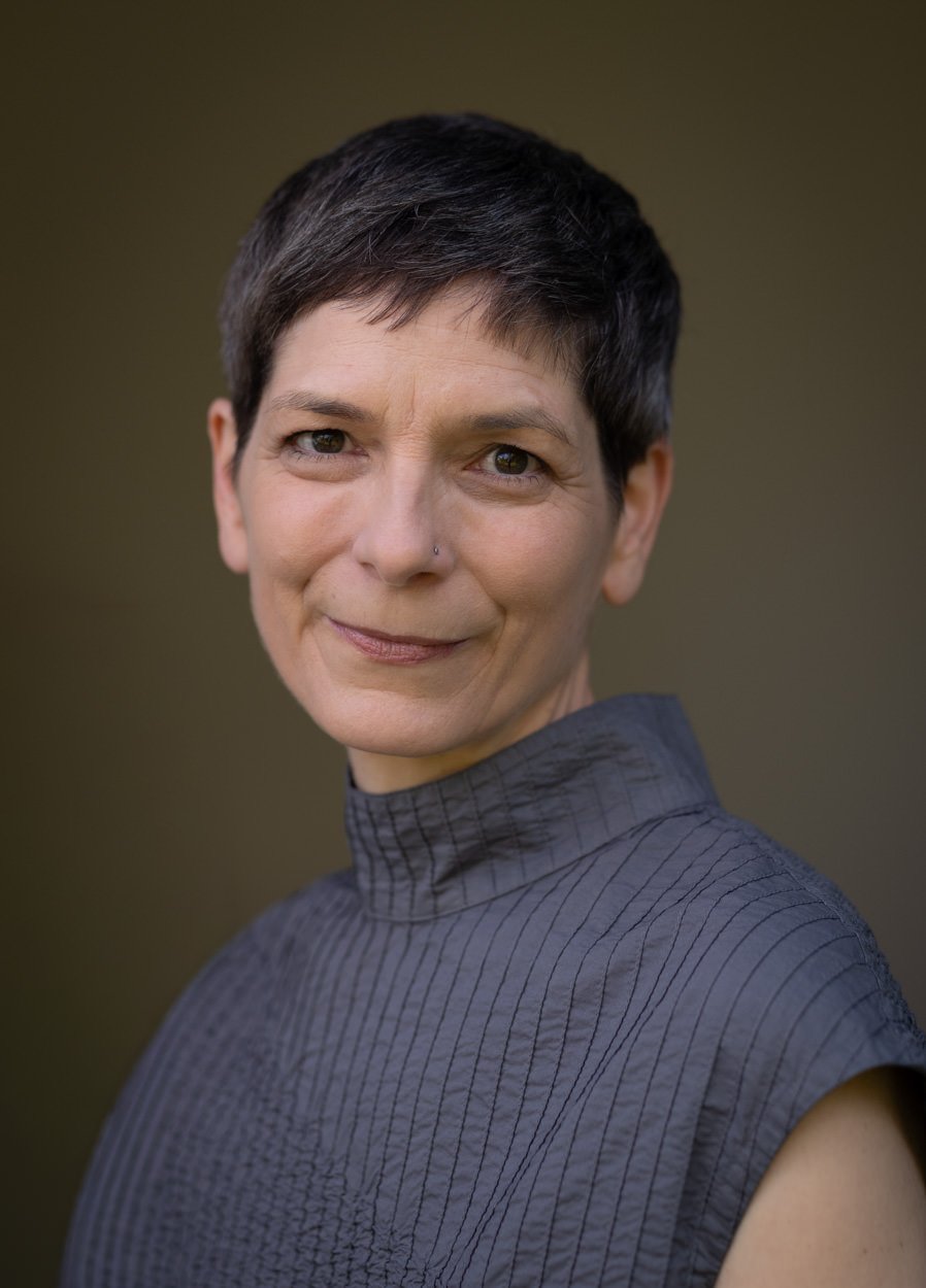 A woman with short dark hair, wearing a gray high-neck sleeveless shirt, looking at the camera with a slight smile against a plain dark background.
