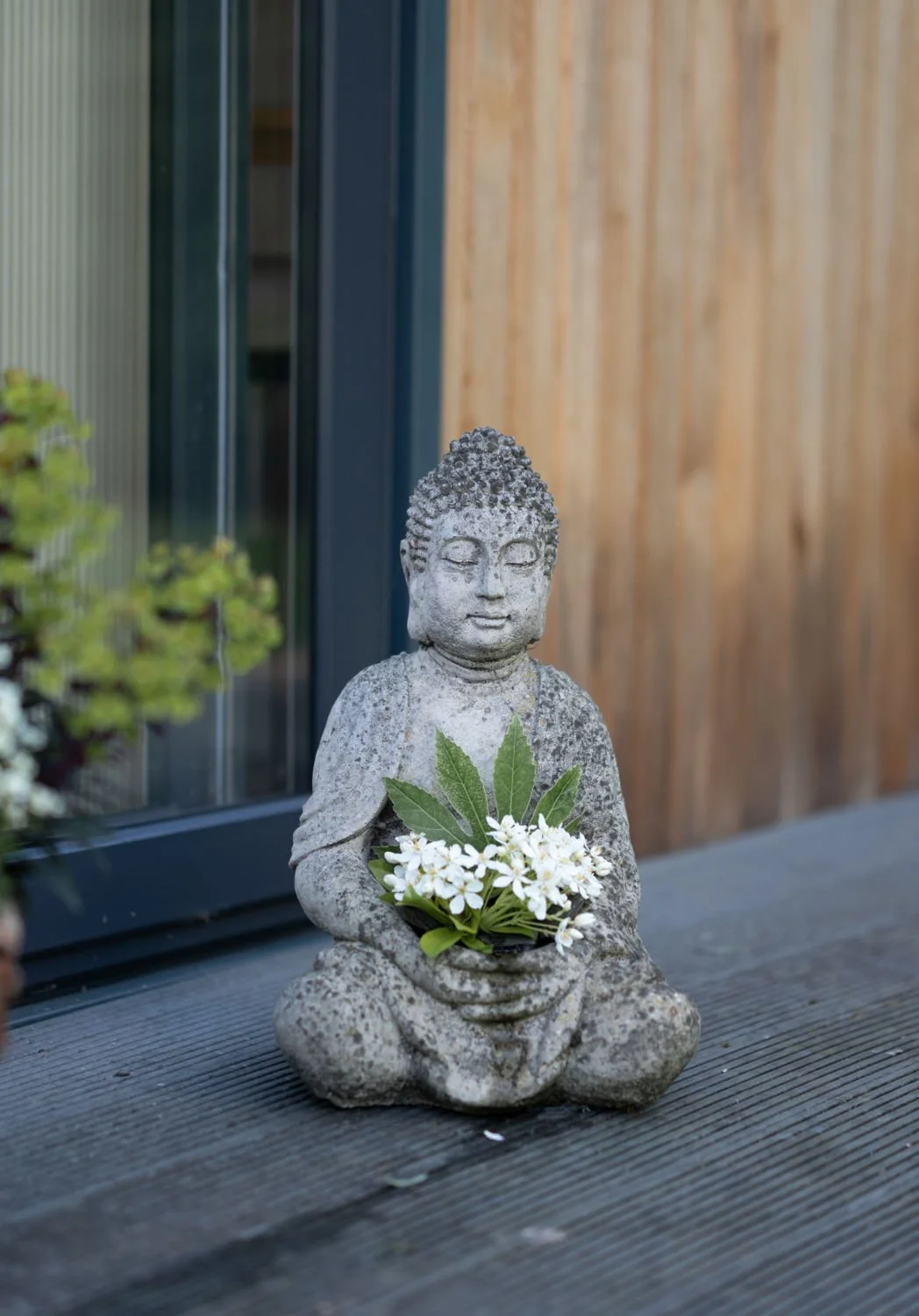 Stone Buddha statue sitting on wooden surface, holding a bouquet of white flowers and green leaves, outside Lethcworth acupuncture premises