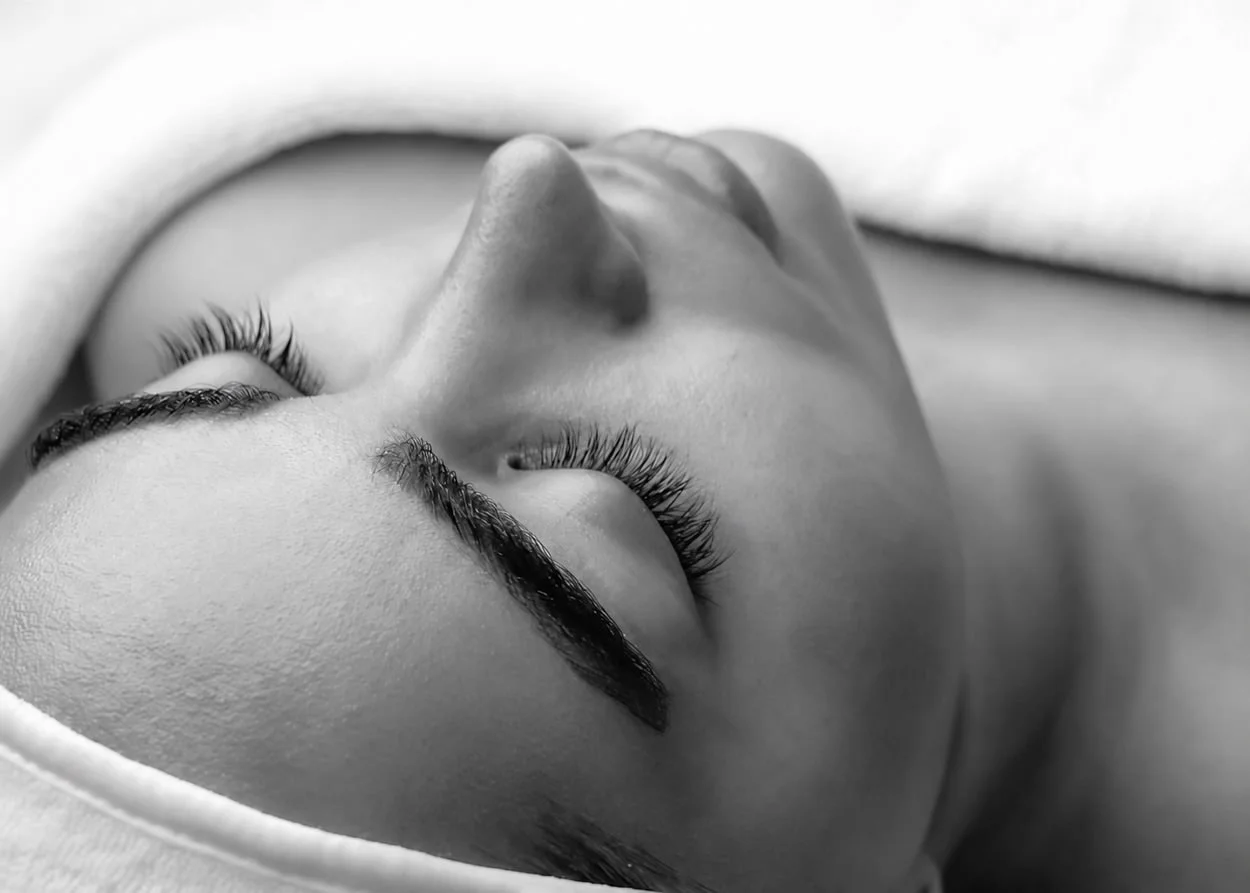 Close-up black and white photo of a woman with long eyelashes, lying down with her eyes closed, under a towel having a facial