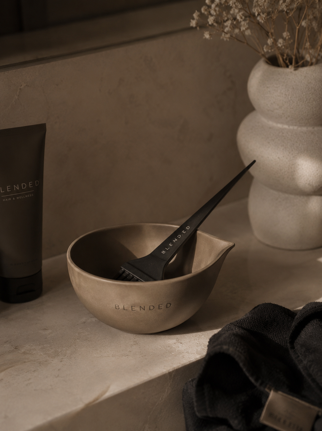 A hairbrush inside a metallic bowl, with the word "BLENDED" on it, on a light-colored surface, beside a black tube of hair product, a dried flower arrangement in a textured ceramic vase, and a folded dark towel.