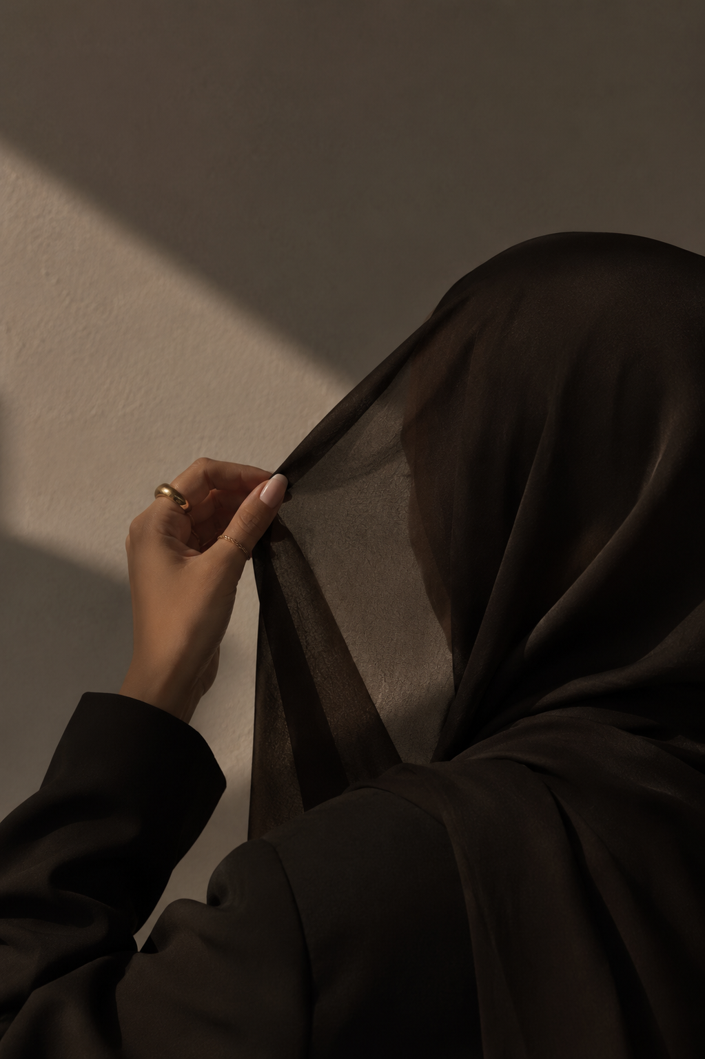 Close-up of a person's hand holding a sheer black fabric, with the person dressed in black, against a neutral wall with light and shadow.