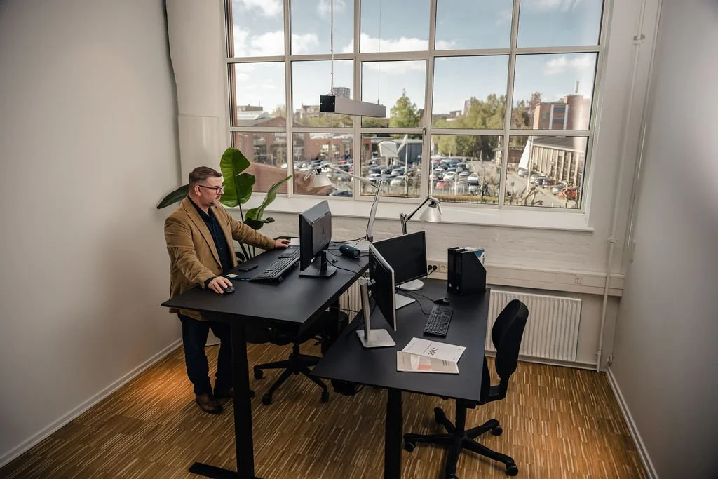 En mand sidder ved et kontorbord med tre computere i et lyst rum med store vinduer og en plante i baggrunden.