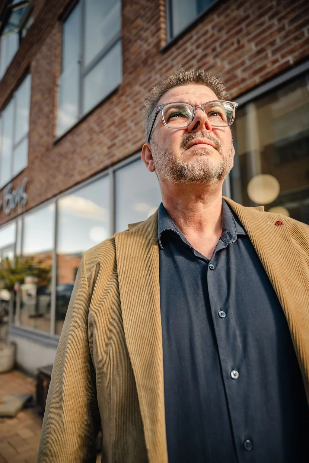 En mand med glaserede briller, gråt skæg og en skjorte i mørkeblå er ude foran en rødbrun murbygning med store vinduer.