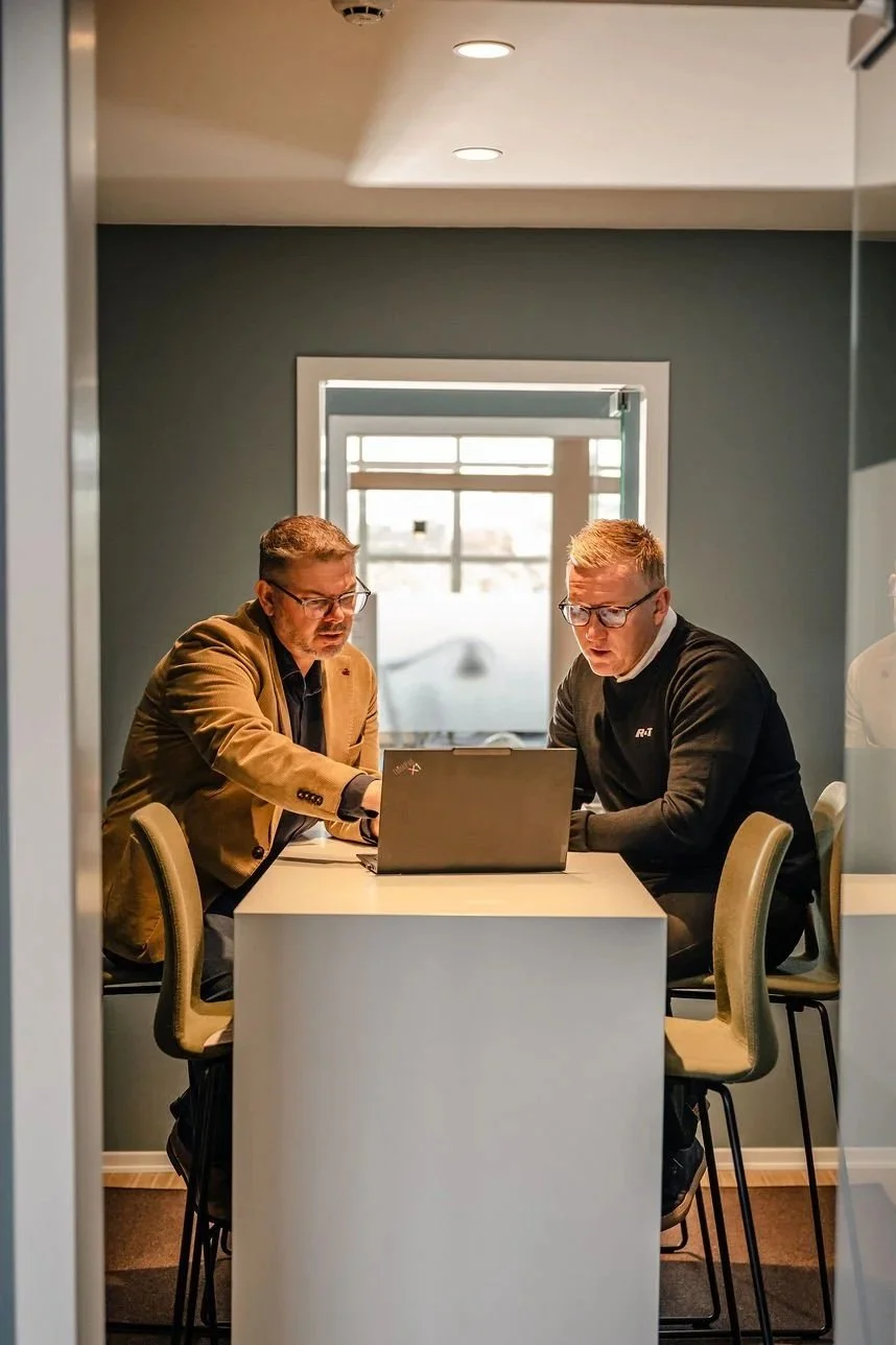 To mænd sidder ved et toiletbord med en bærbar computer i en skærm, indendørs med en vindue i baggrunden.