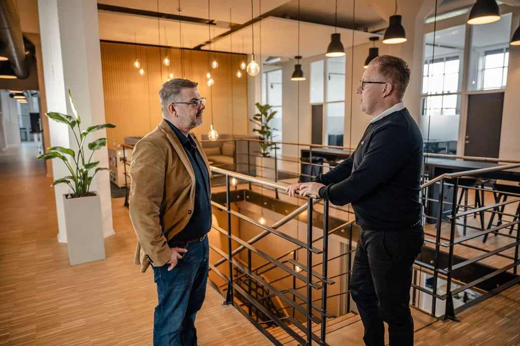 To mænd taler på en altan i et moderne kontormiljø med træbord og grønne planter.
