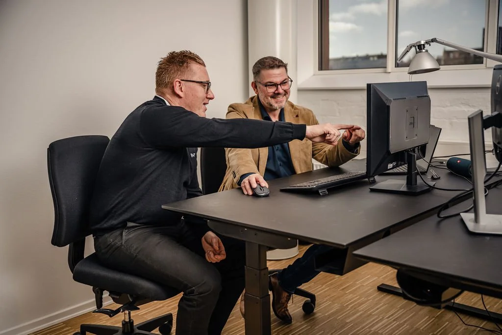 To mænd, der sidder ved et skrivebord med en computer, de ser glade ud og peger på skærmen.