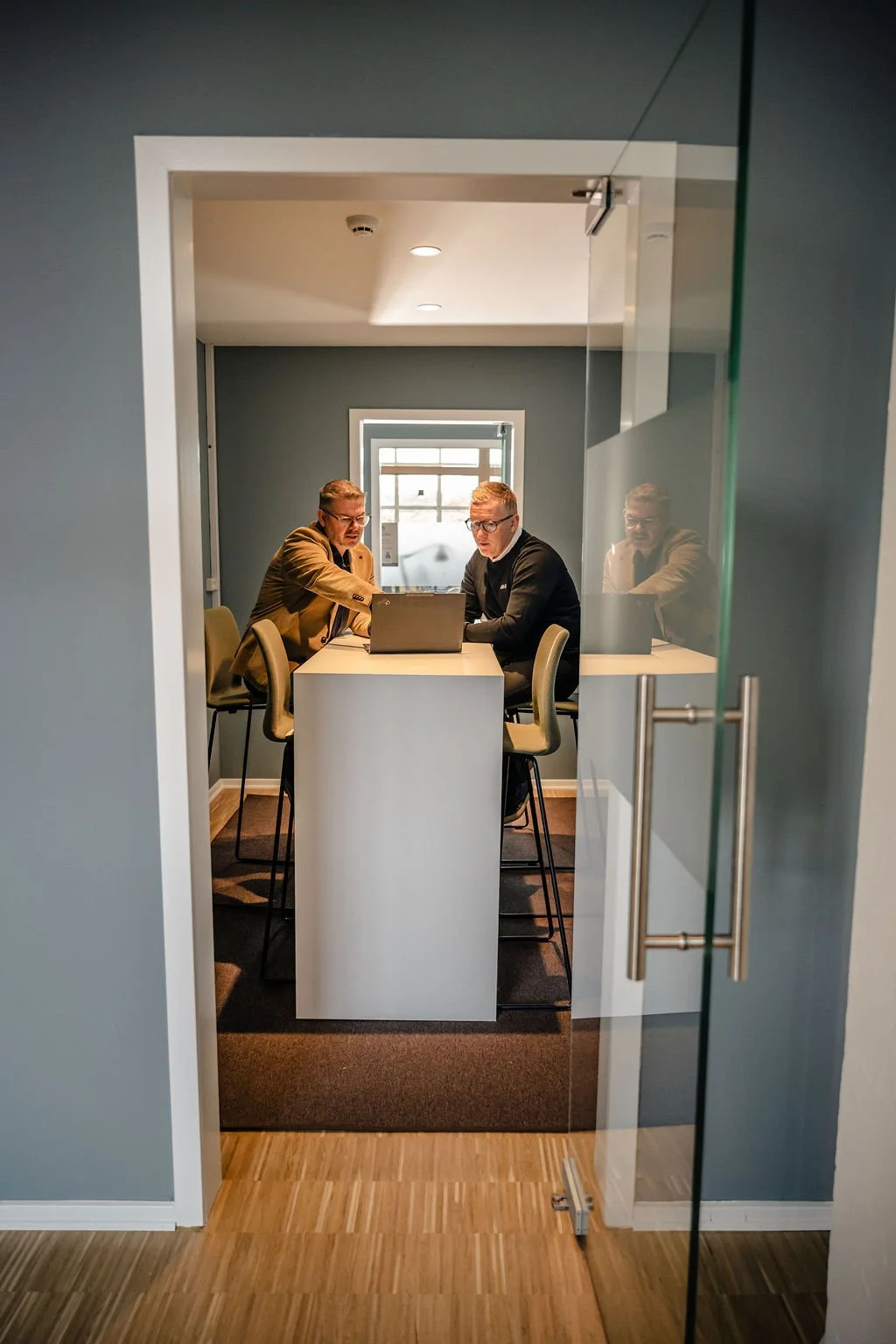 To mænd sidder ved et bord med bærbare computere i en lille mødelokale set gennem en glasdør.