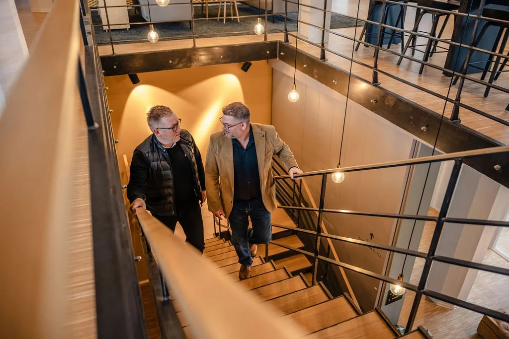 To mænd går op ad en trappe i et moderne indendørs område med trætrin, sorte gelændere og varme belysningslamper.