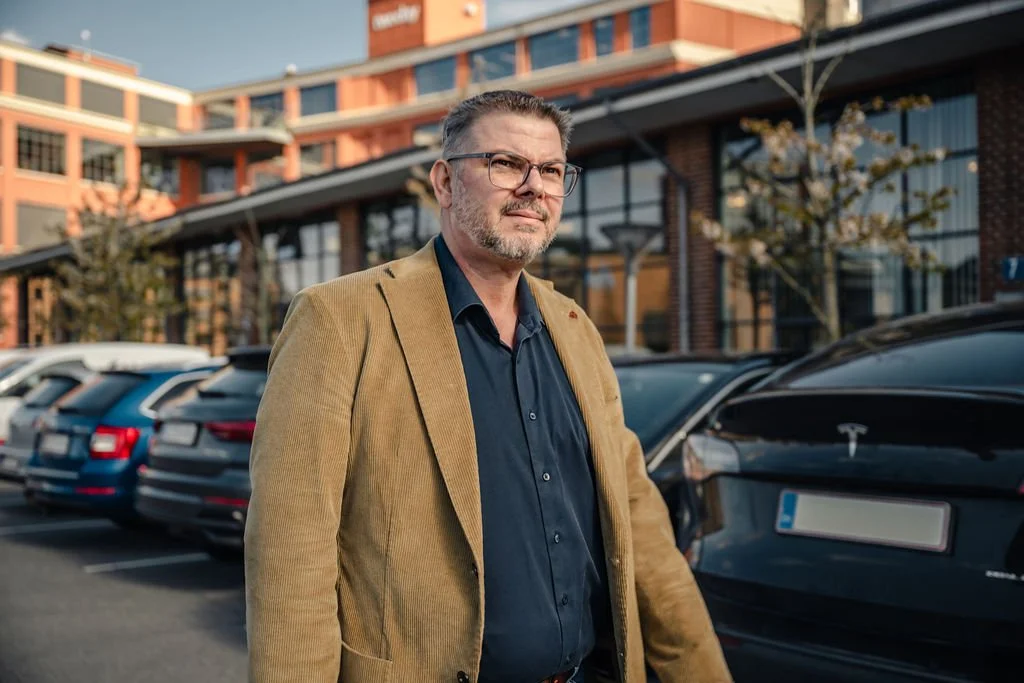 En mand med briller og gråt skæg står foran en parkeringsplads med flere biler og moderne bygninger i baggrunden.