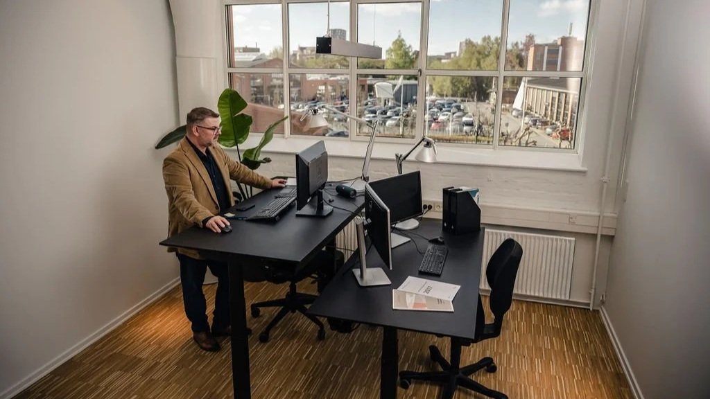 Mand sidder ved en computer i et kontormiljø med store vinduer og udsigt over parkeringsplads og bygninger.
