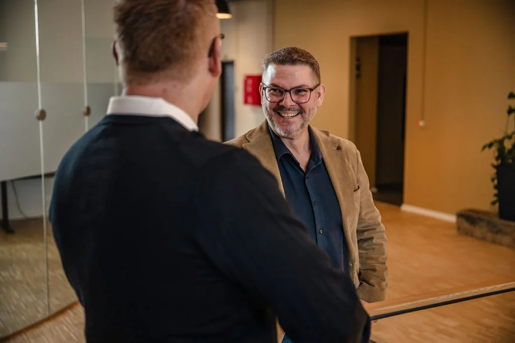 To mænd taler med hinanden i et kontormiljø, smilende og i samtale.