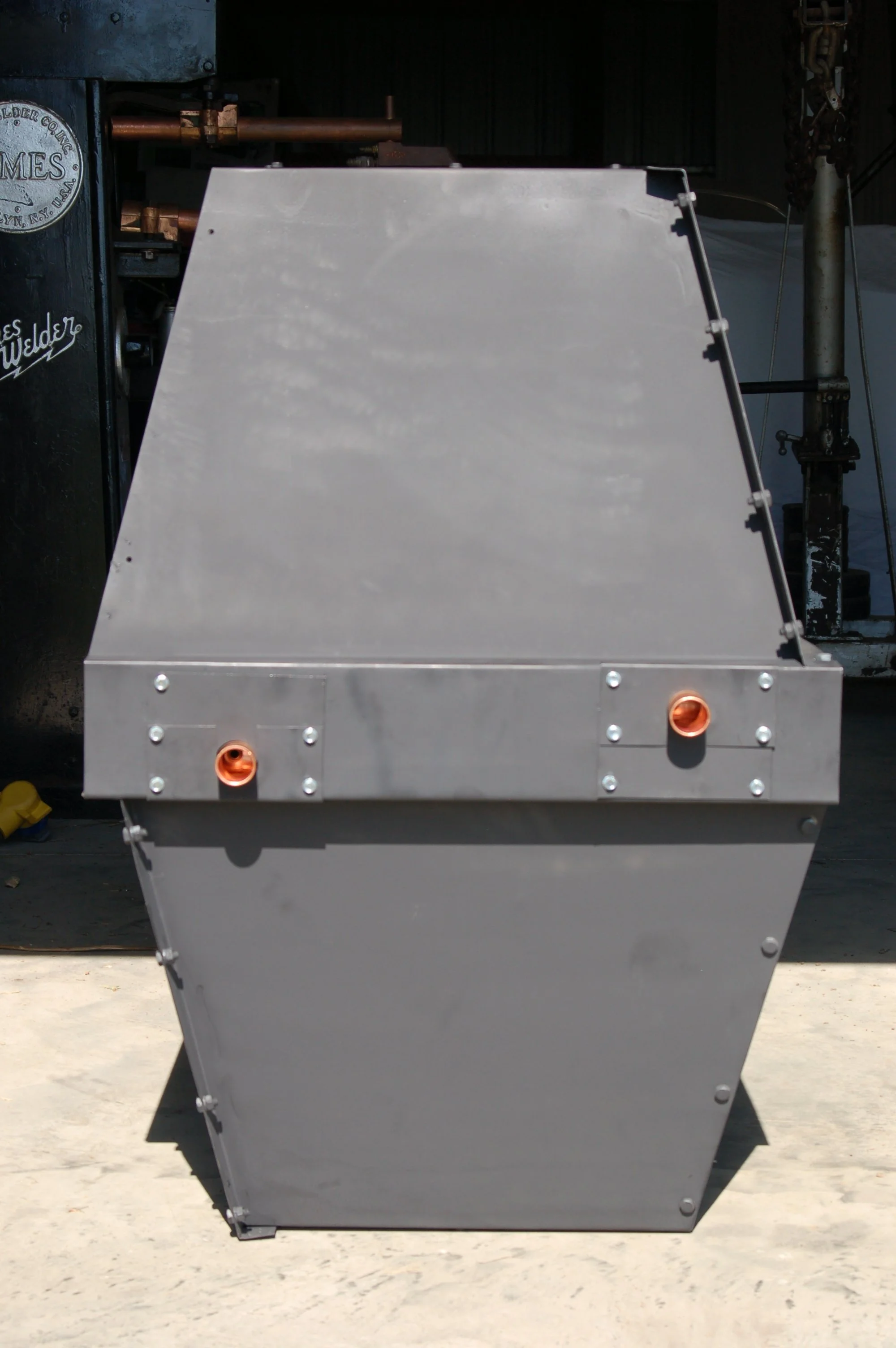 Large gray industrial machine or container with mechanical components. It has two copper pipes, orange openings, and secured bolts, set on a concrete surface with a dark background.