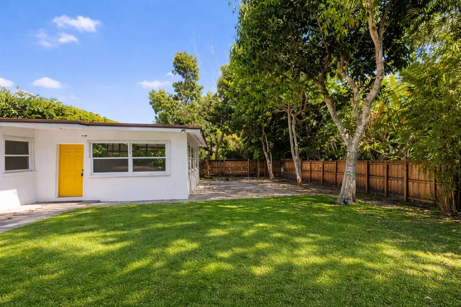 A backyard with green grass, several mature trees, a white house with yellow door and large windows, a stone pathway, and a wooden fence under a blue sky with some clouds.