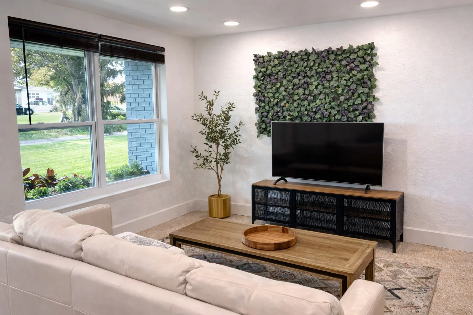 Living room with large window, white sofa, wooden coffee table with tray, TV on a black and wood stand, green wall art, and a potted plant, outside view of greenery and houses.