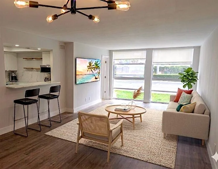 Modern living room with a white sofa, a round coffee table, a beige armchair, and a large window with blinds. There is a green plant, colorful pillows, and a flat-screen TV on the wall. A chandelier hangs from the ceiling, and the space is connected 