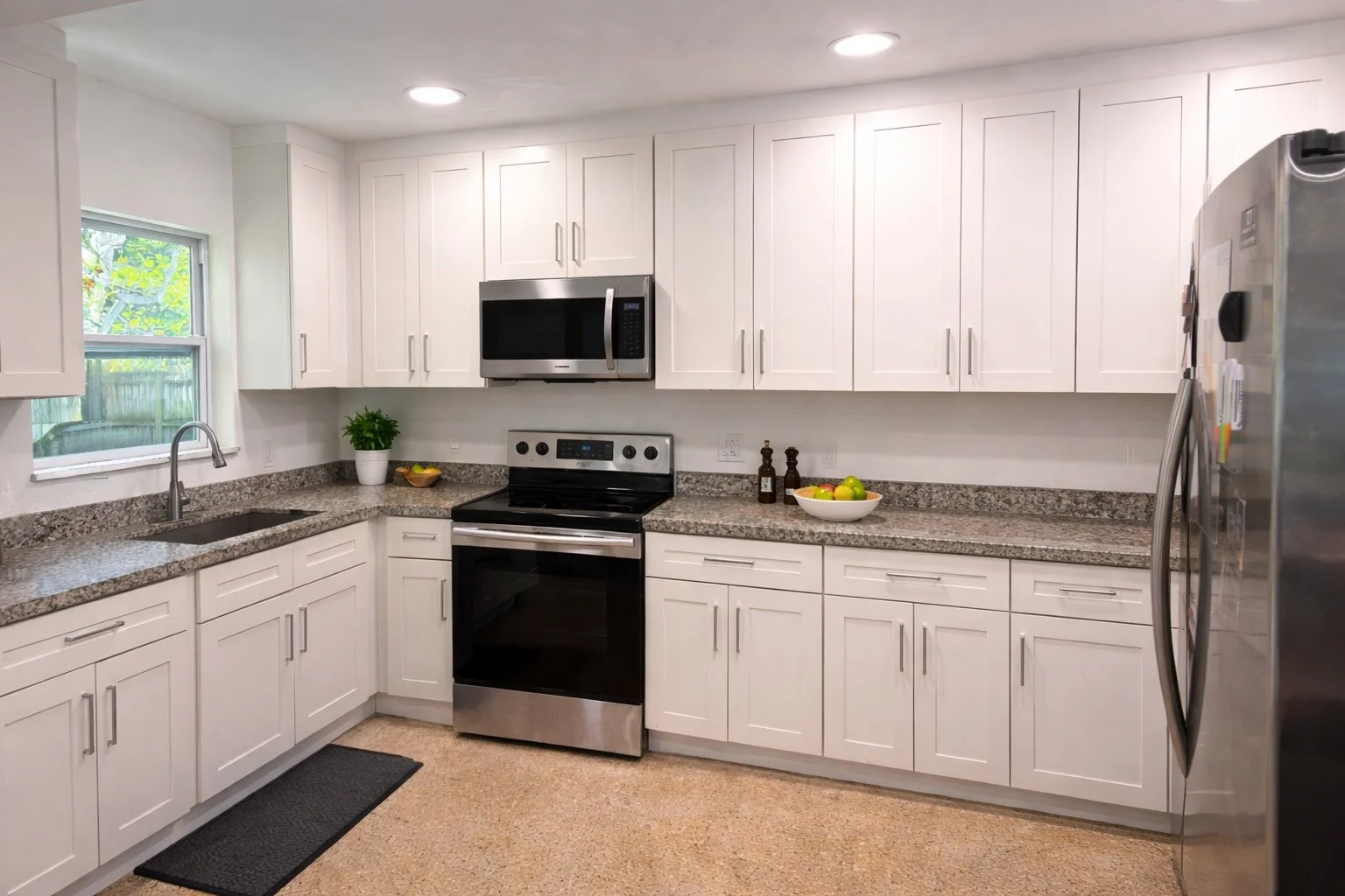 Modern kitchen with white cabinets, granite countertops, stainless steel appliances including a microwave, oven, and refrigerator, and a window above the sink with a view of greenery outside.