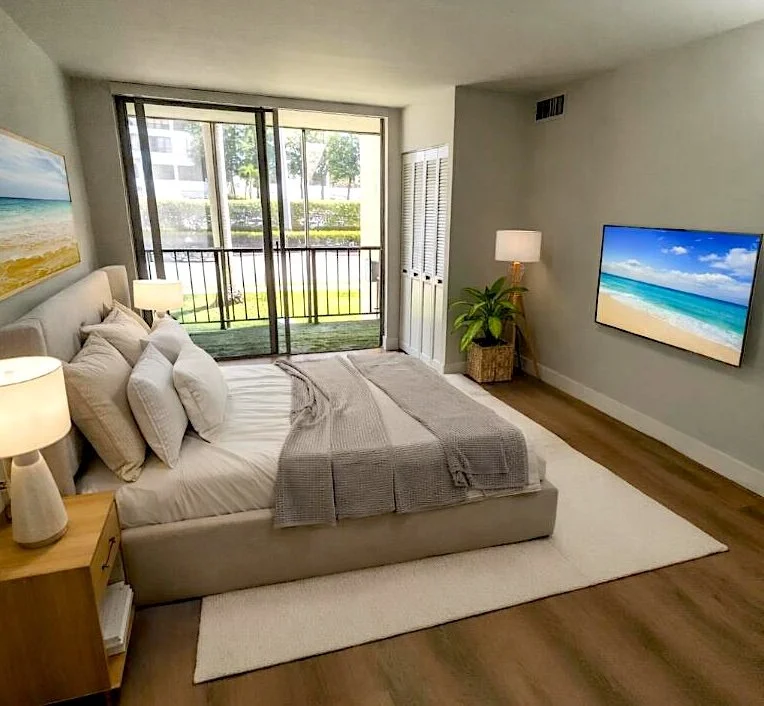 Modern bedroom with a large bed, bedside lamps, a flat-screen TV showing a beach scene, a potted plant, and sliding glass doors leading to a balcony with a view of trees and buildings.