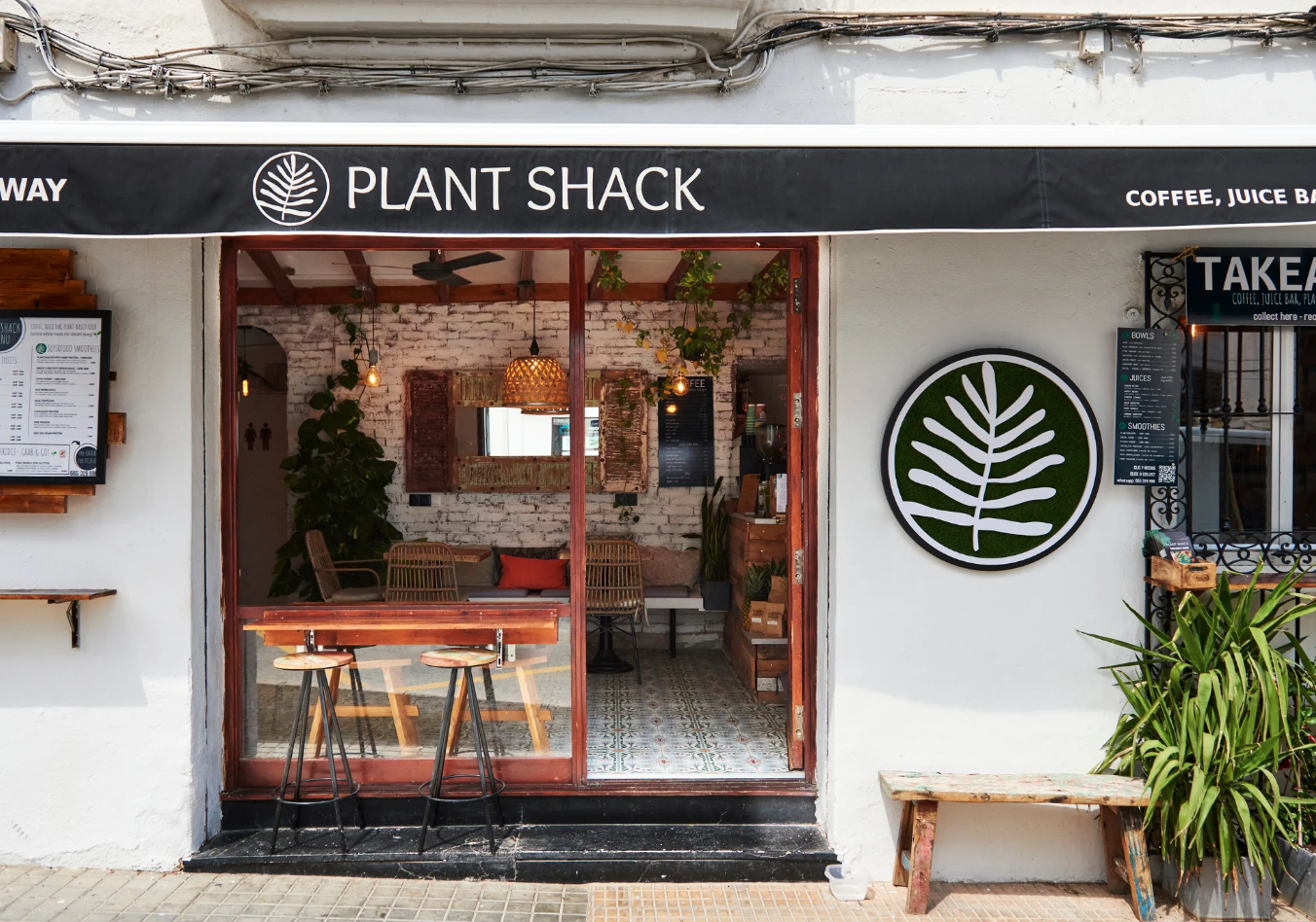 Exterior of the first Plant Shack in Altea, Alicante, Spain with an open entrance, a black awning with white text and logo, and interior seating with wooden chairs and plants.