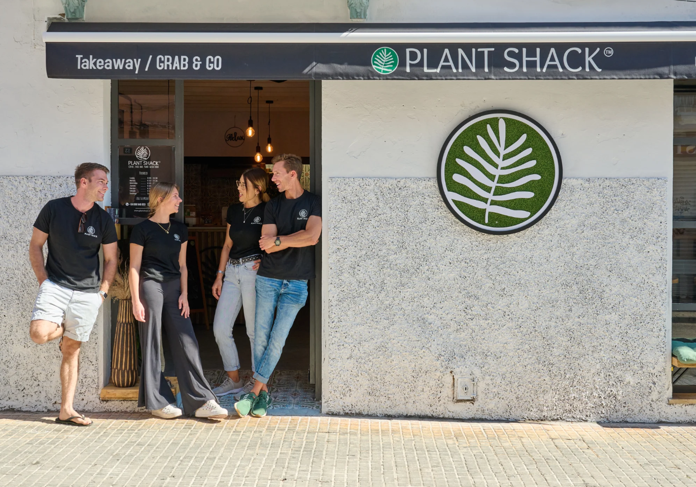 Four people standing outside a Plant Shack restaurant, smiling and talking, with black shirts and a black awning with white text.