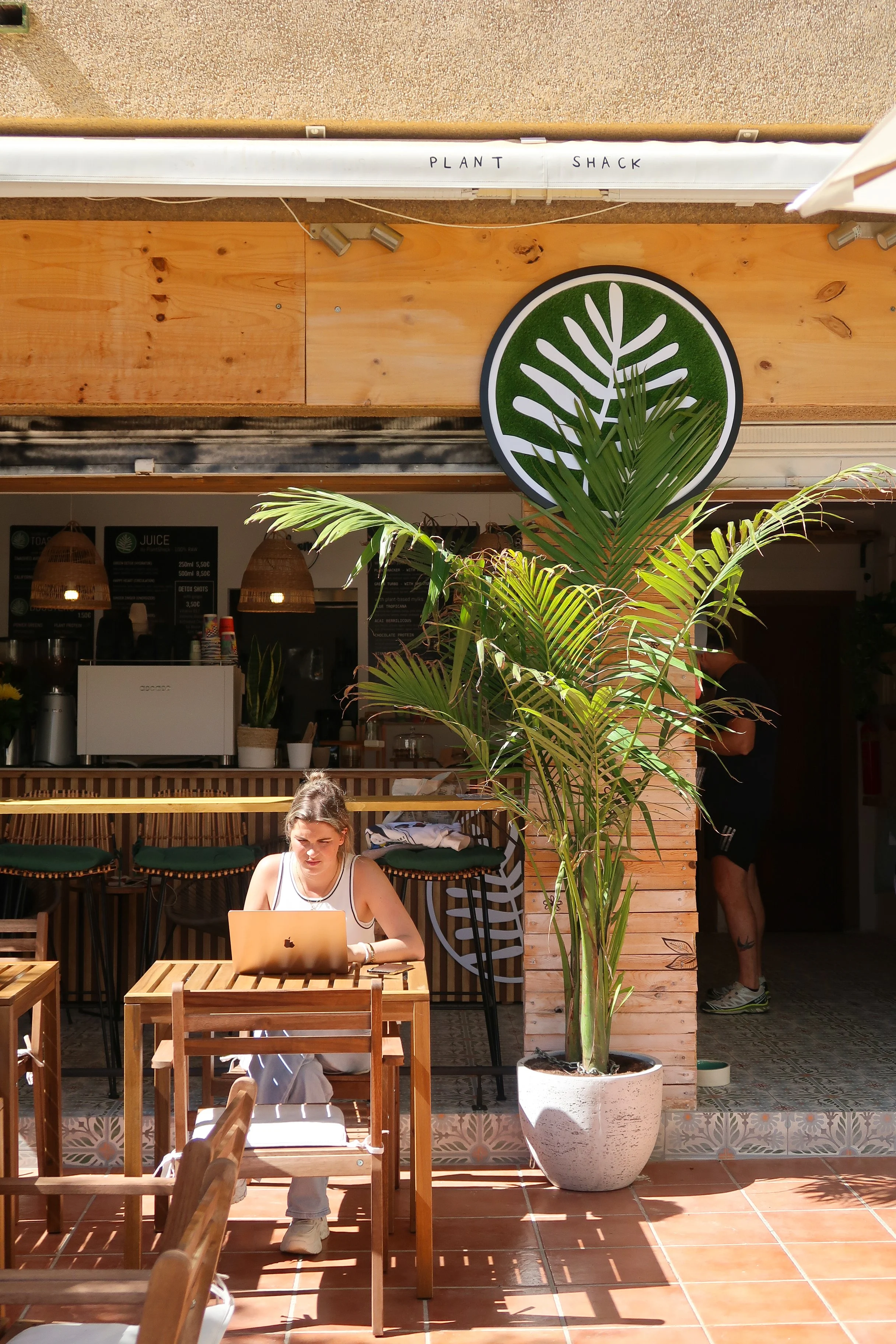 Woman sitting at a wooden table outside a Plant Shack working on a laptop, with large potted palm plant nearby and a sign reading 'Plant Shack' overhead.