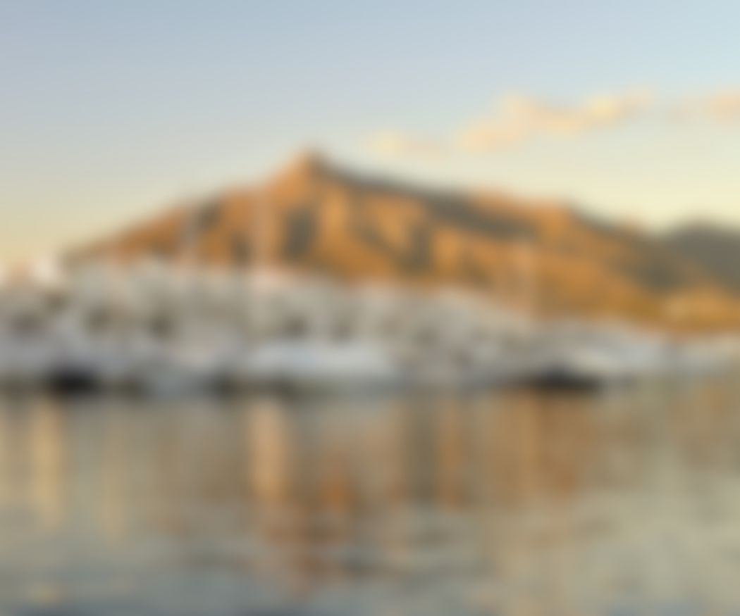 A blurred photo of a mountain with a building on top, surrounded by water with boats docked nearby during sunset.