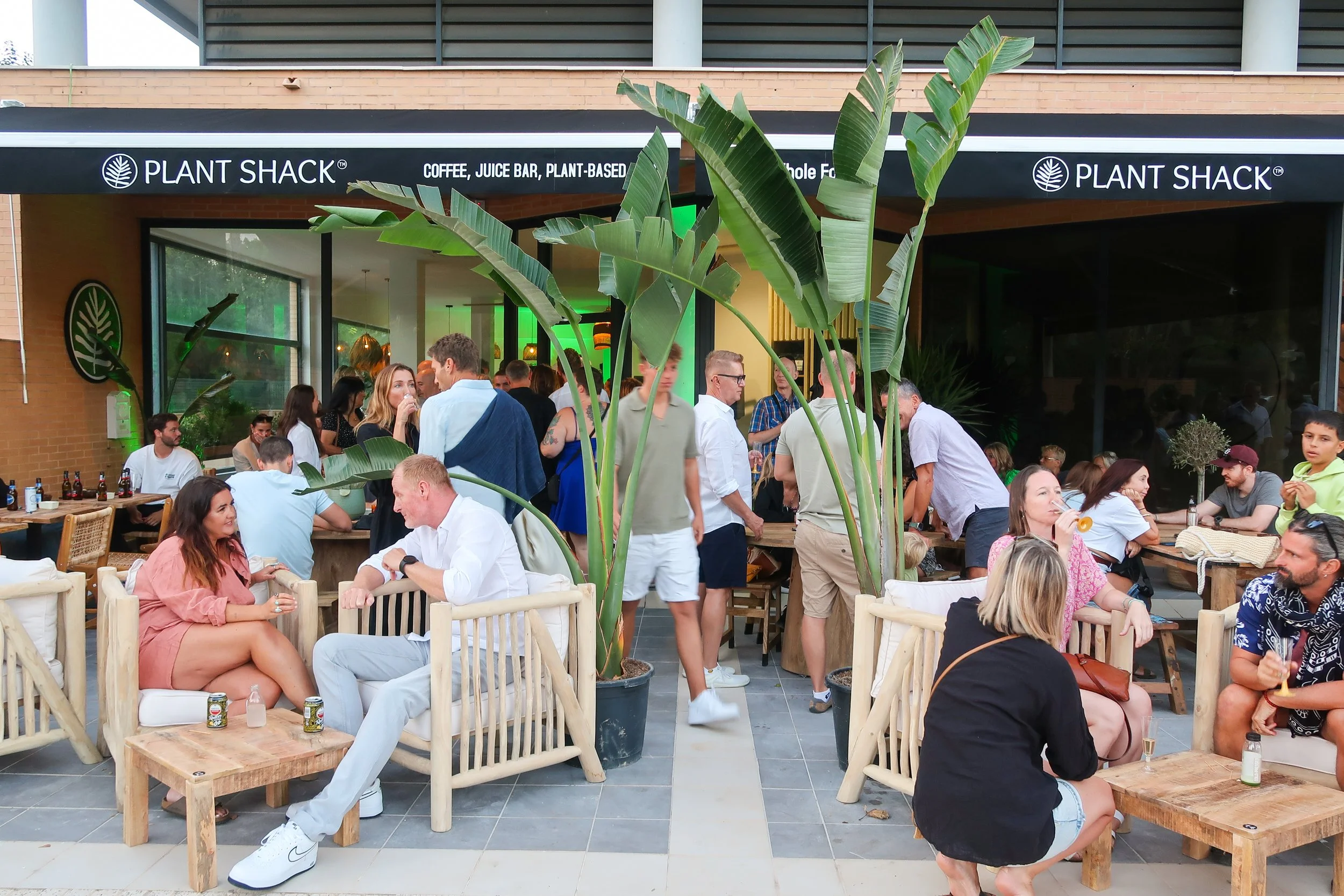 People socializing outside a Plant Shack restaurant with a drink in hand, tall green plants in the foreground, and a modern building in the background.