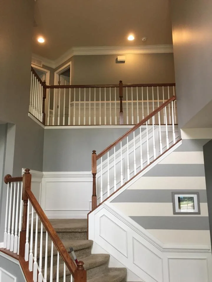 Interior view of a staircase with beige carpet, white and wood handrail, light grey walls, and a second floor balcony with white railing. Small window on the wall near staircase.