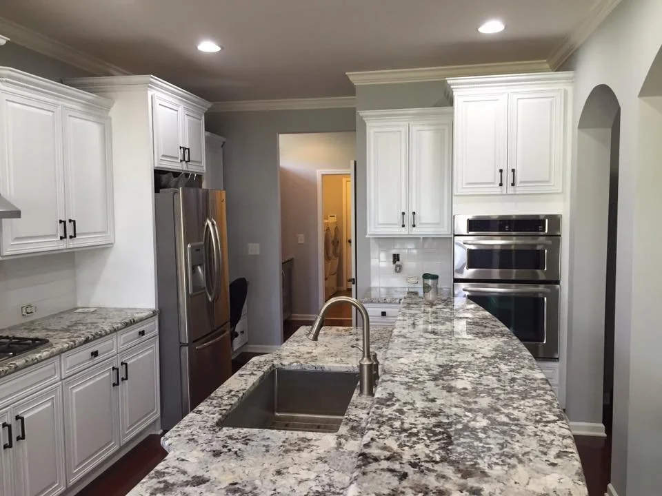 Modern kitchen with white cabinets, granite countertops, stainless steel appliances including a refrigerator and double oven, and a curved kitchen island with a sink.