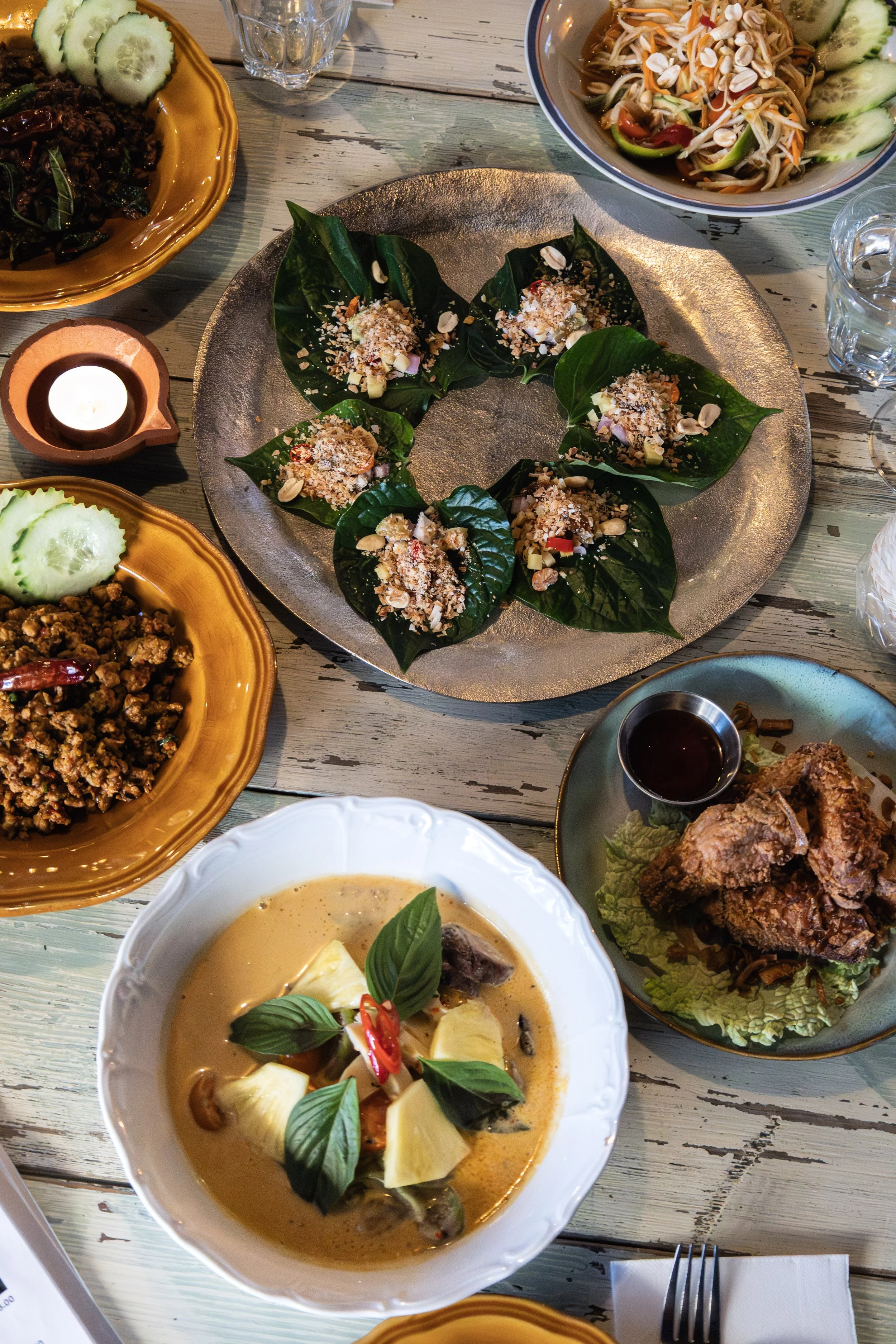 Un repas avec plusieurs plats thaïlandais, comprenant desnouvhées thaies au feuilles de bétel, un curry rouge au magret de canard, ananas frais pousses bambou et aubergines thaïes et enfin le Pad Kaprao, un sauté de boeuf au basilic thaï et piments