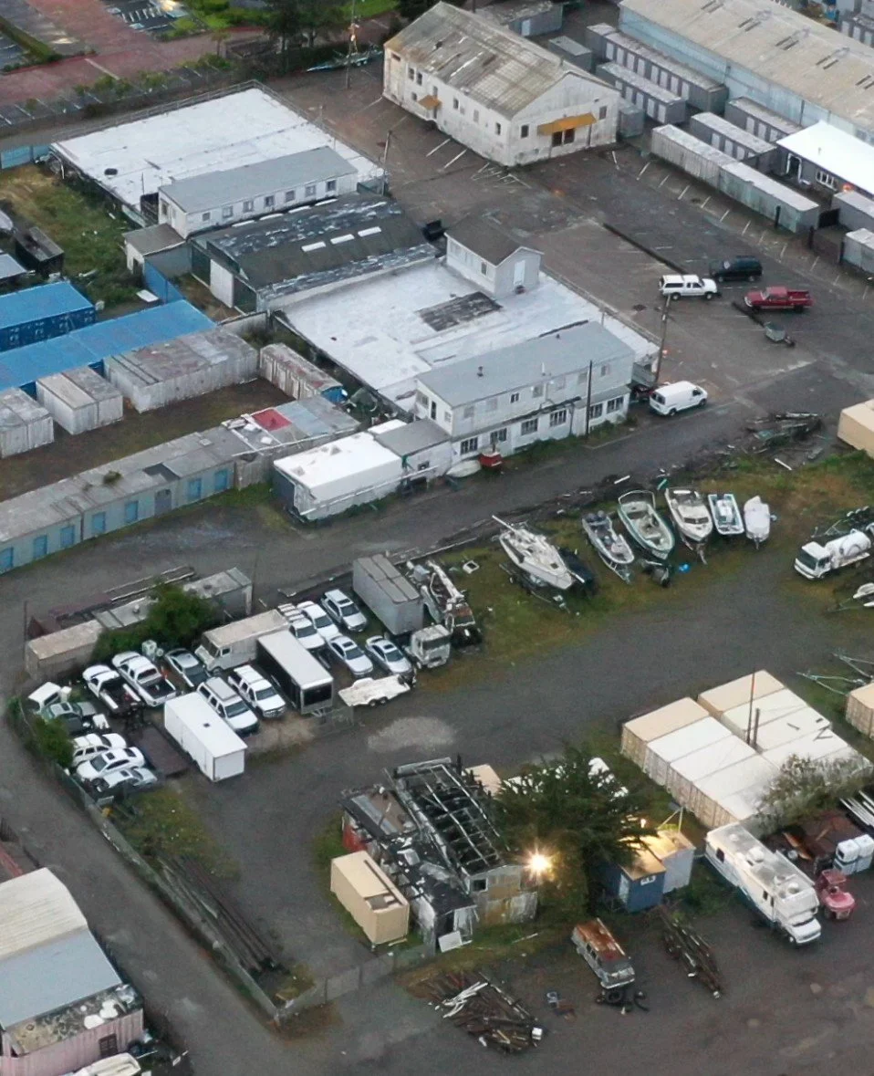 Aerial view of Sausalito Waterfront & Industrial zone lot with damaged and intact vehicles, some boats, and small structures, including a heavily burned building and parking areas.
