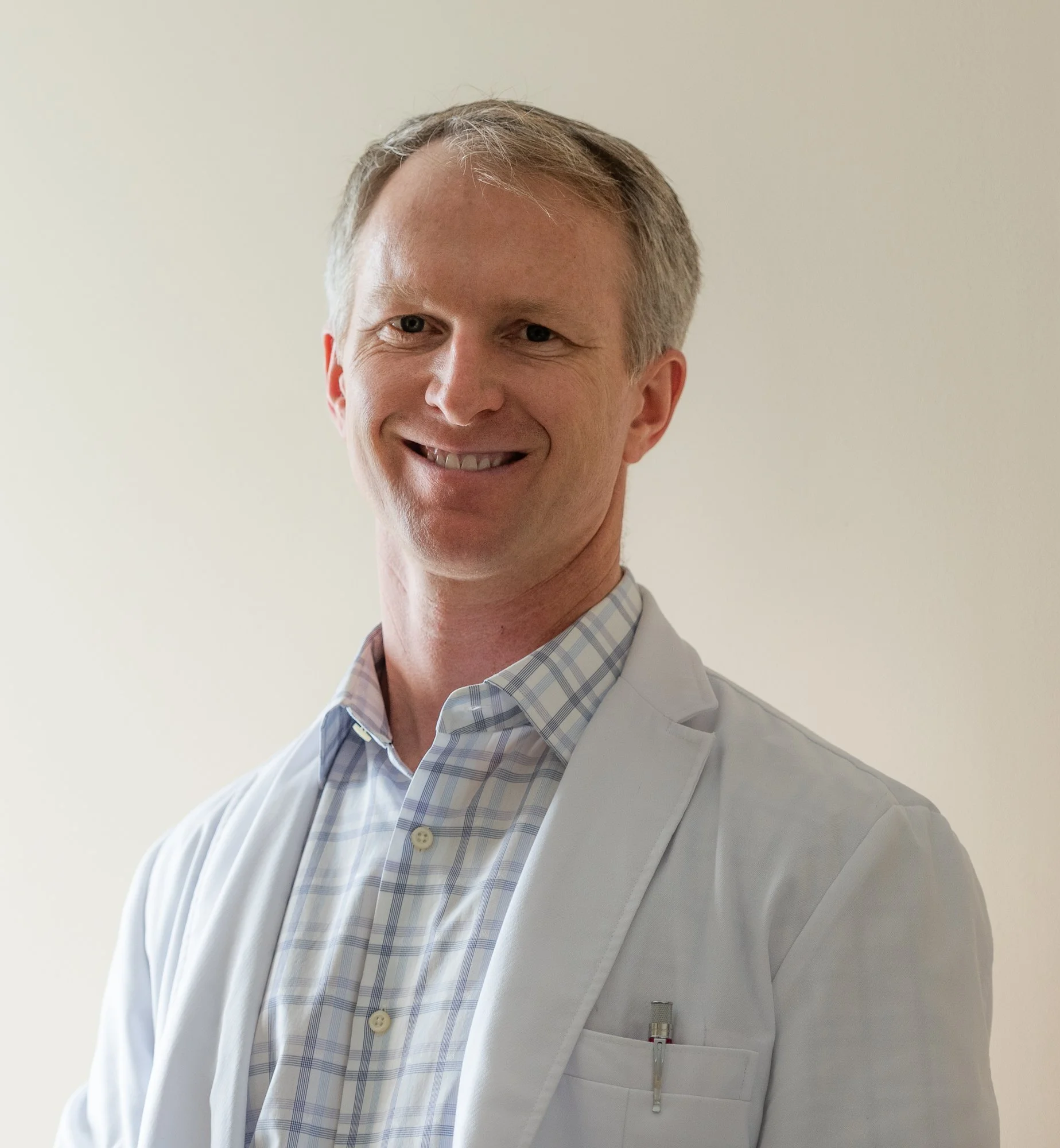 A smiling male doctor in a white coat and plaid shirt, with a pen in his pocket, standing against a plain light-colored background.