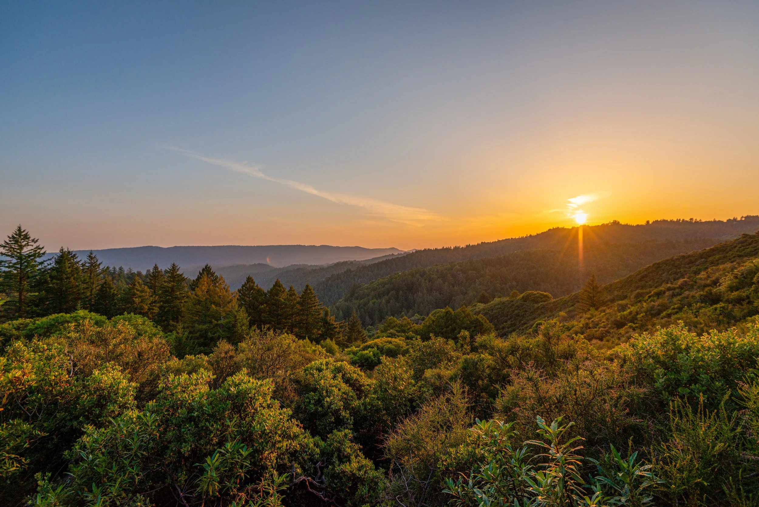 Sunset over a forested mountain landscape with a clear sky and a few clouds.