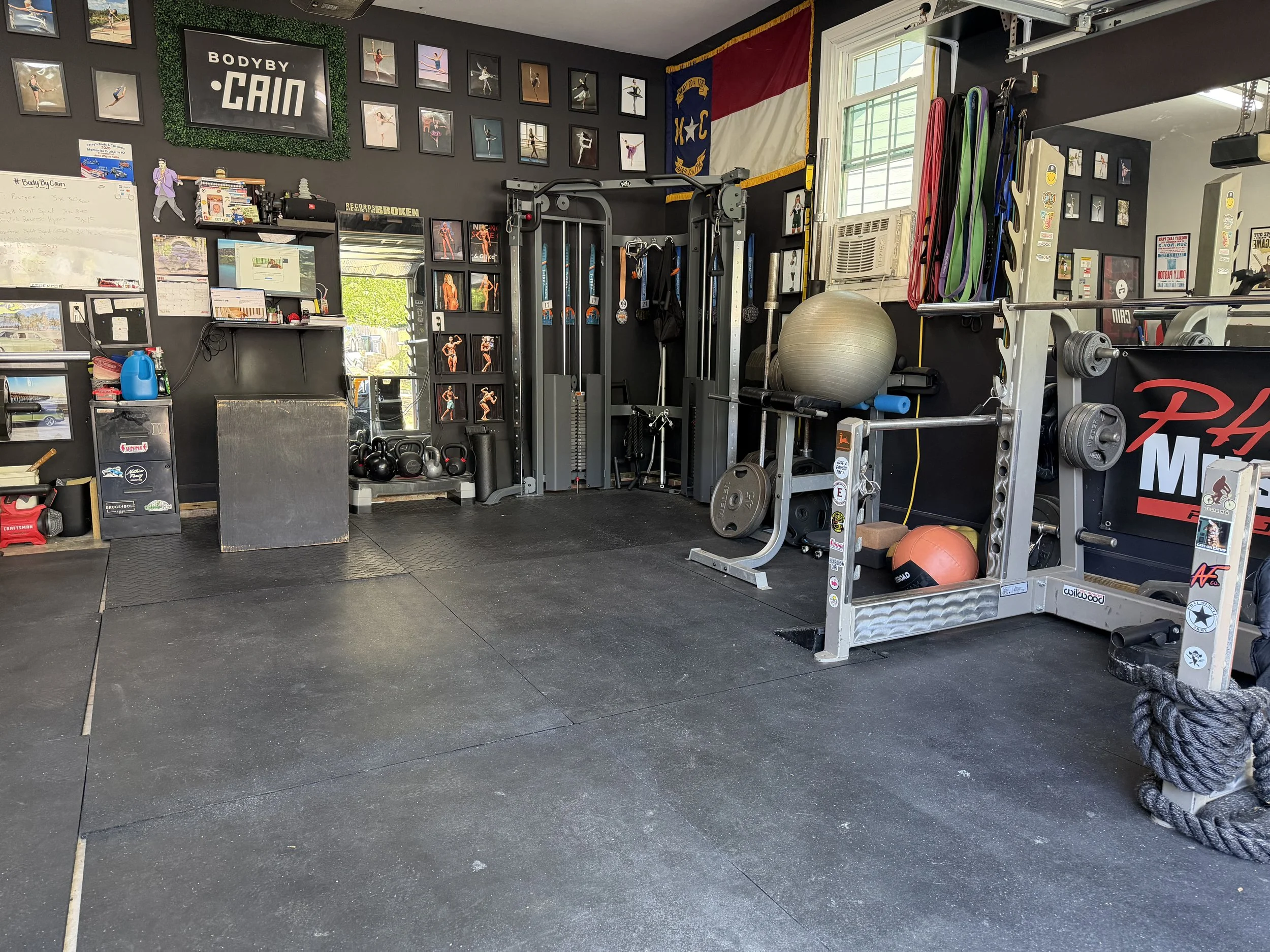 Interior of a gym with exercise equipment, framed photos of ballet dancers on dark walls, a flag, and a window with sunlight.