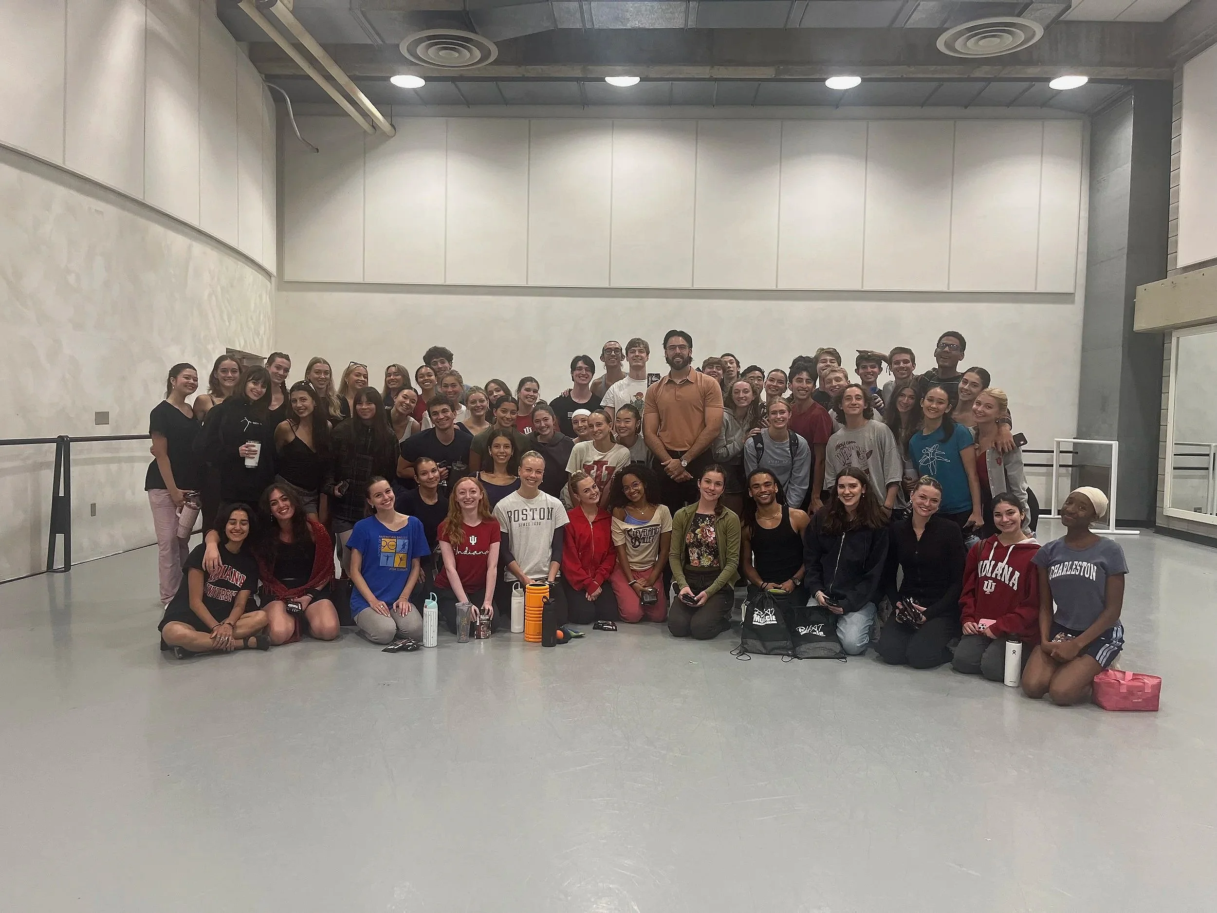 A large group of diverse young people and a few adults gathered together for a group photo inside a spacious room with white walls and a gray floor. The group is smiling and casually dressed, some holding water bottles and bags, with a few standing and some sitting or kneeling in front.