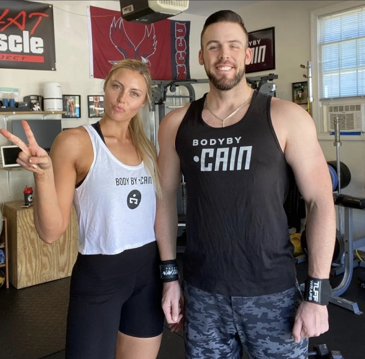 A woman and a man in a gym posing for a photo. The woman makes a peace sign and wears a white tank top and black shorts. The man wears a black tank top and camouflage shorts. Both are smiling and wearing wristbands.